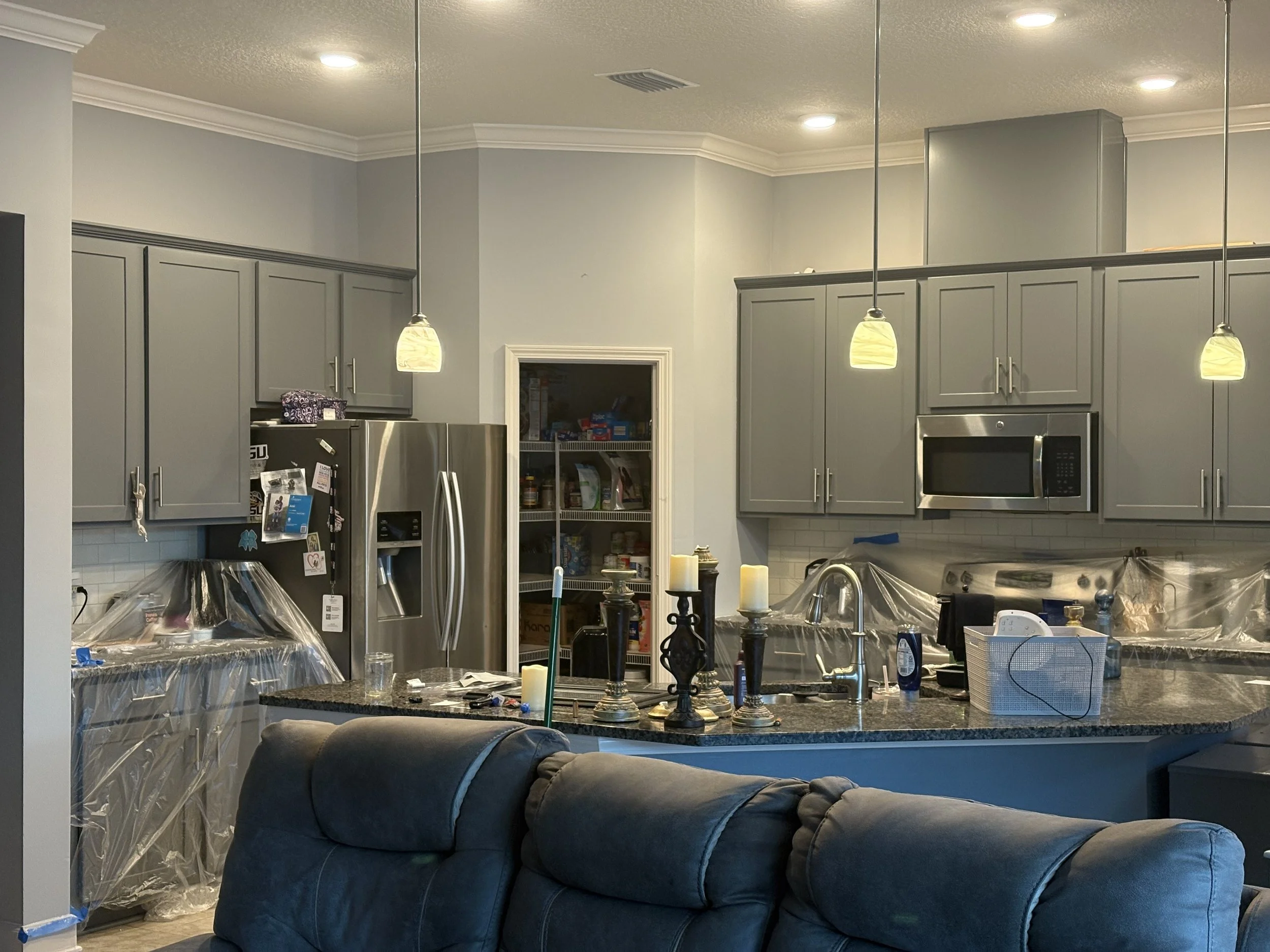 A kitchen with gray cabinets, a granite countertop, a stainless steel refrigerator, and a microwave. There are pendant lights hanging from the ceiling, and the kitchen appears to be under renovation or decoration, with plastic coverings and various items on the counter.