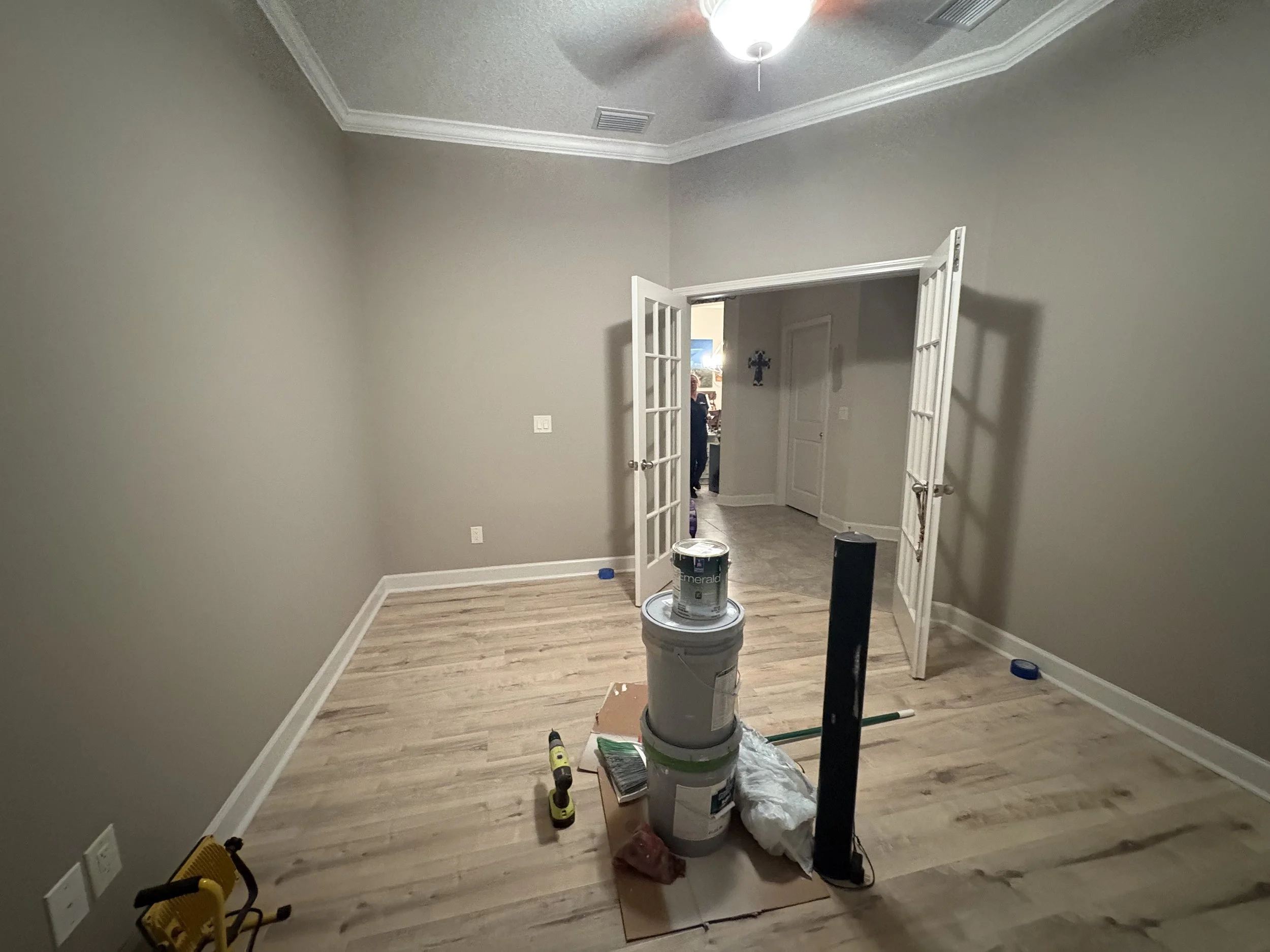 Room renovation in progress with painting supplies, paint cans, a cordless drill, a box, and construction tools on a wooden floor, beige walls, and a ceiling fan in the room.