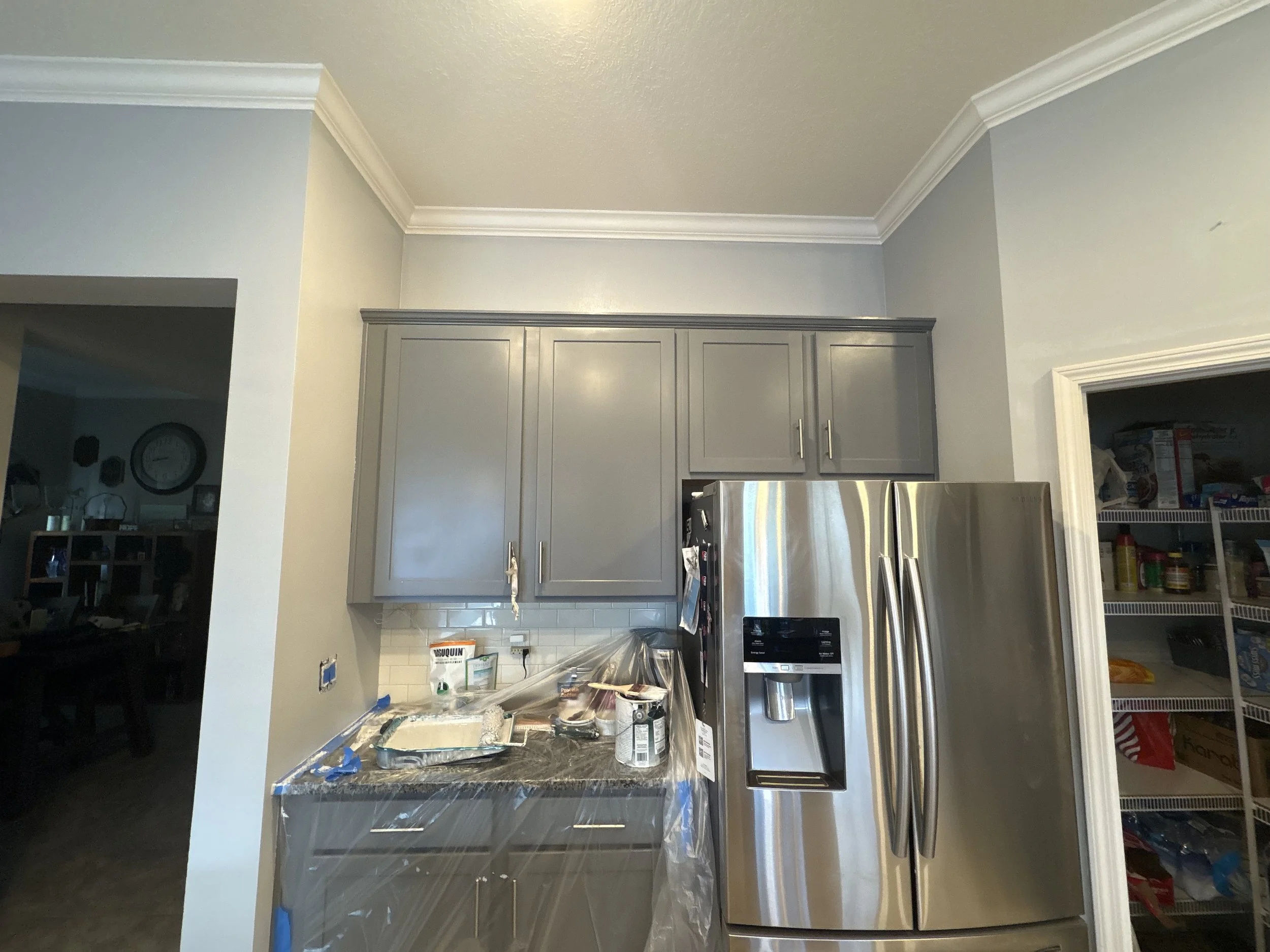The image shows a kitchen with gray cabinets, a stainless steel refrigerator, and a kitchen counter with various food preparation items and ingredients. There is a pantry on the right and a doorway on the left leading to another room.