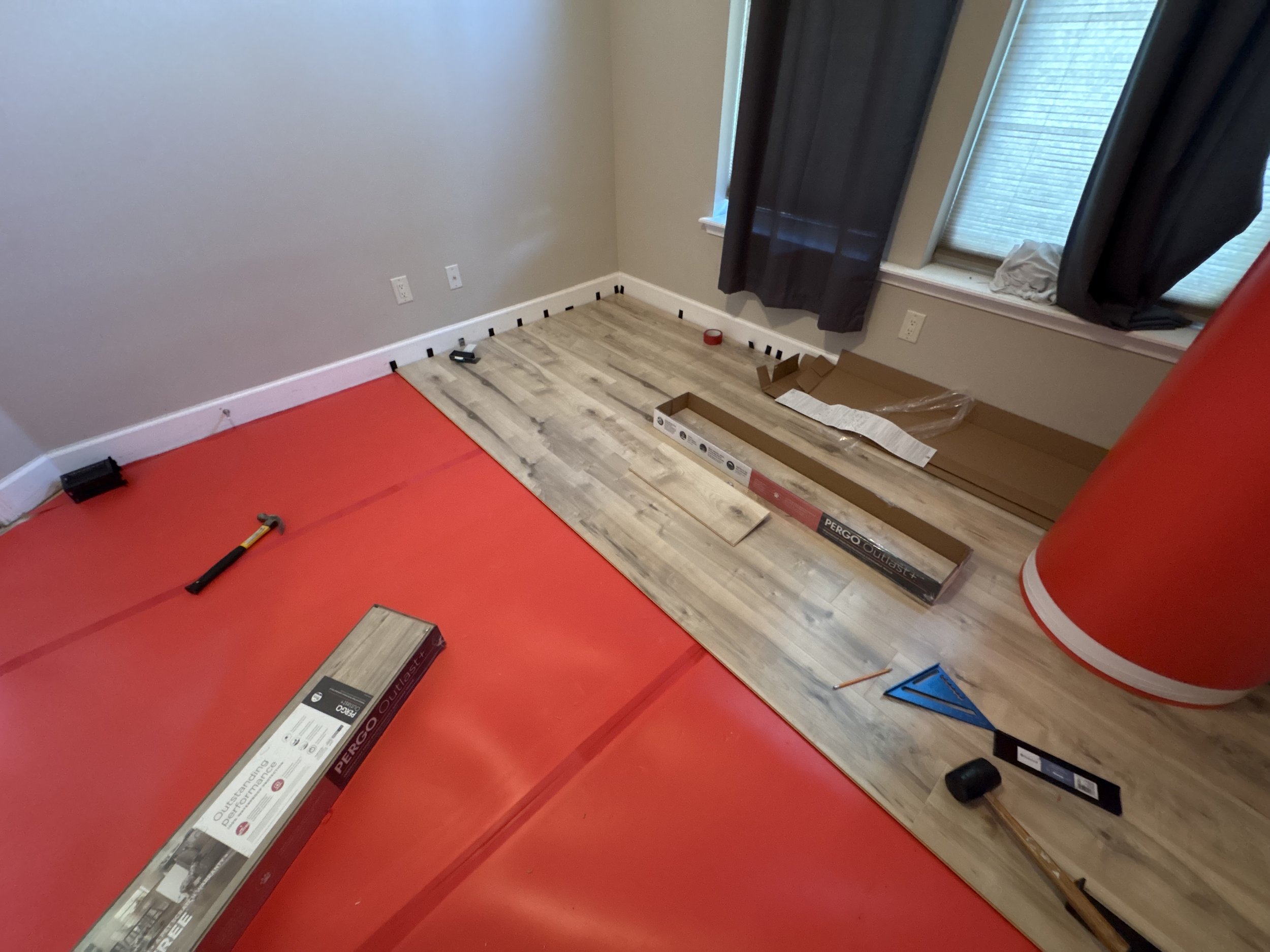 Room with light-colored walls, black checkers along the baseboard, a window with closed black curtains, and flooring partially covered with cardboard and tiles, with a red plastic sheet and tools on the floor.