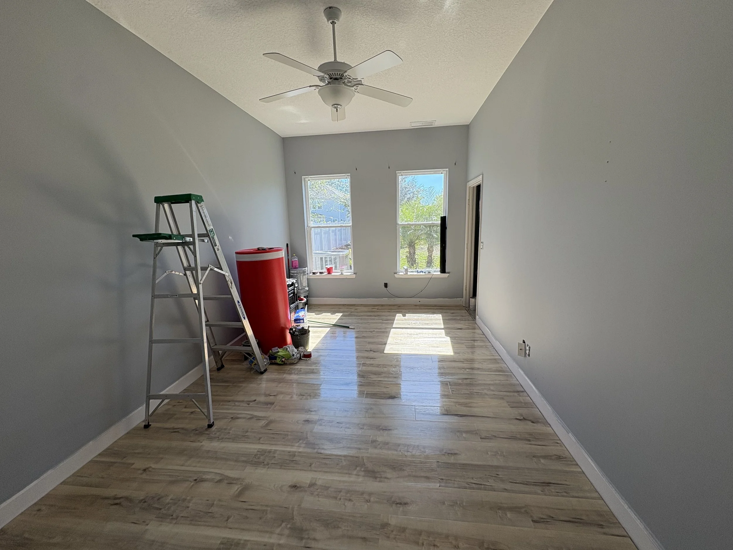 Empty room with gray walls, hardwood floor, three windows, window sill with some items, white ceiling fan, ladder, and construction items.