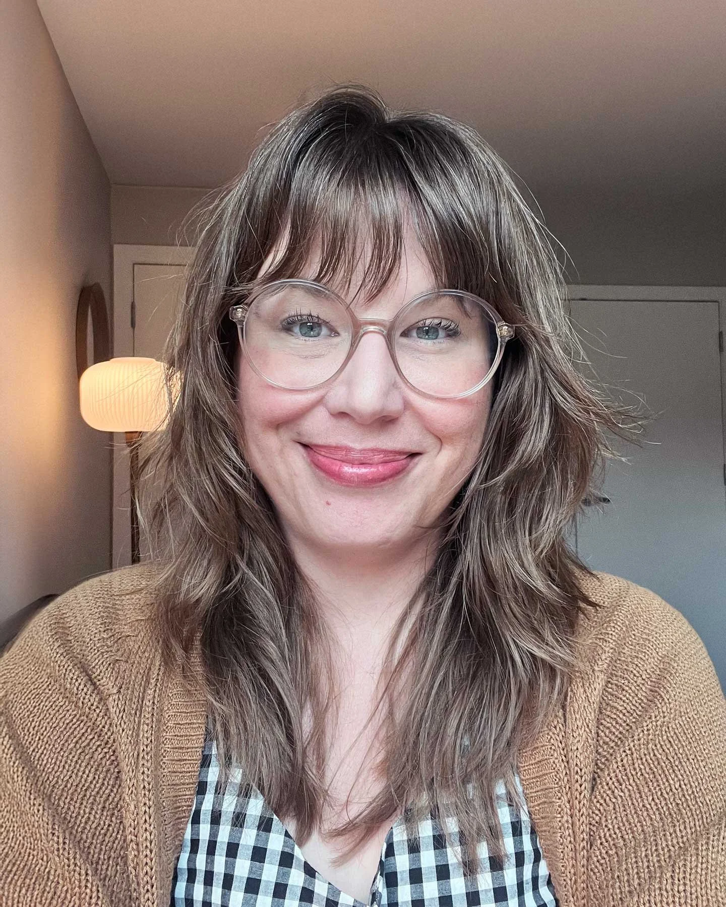 Crystal Tracy with glasses, long wavy brown hair, wearing a brown knit sweater over a black and white plaid shirt, smiling in a room with neutral-colored walls, a white door, and a lamp in the background.