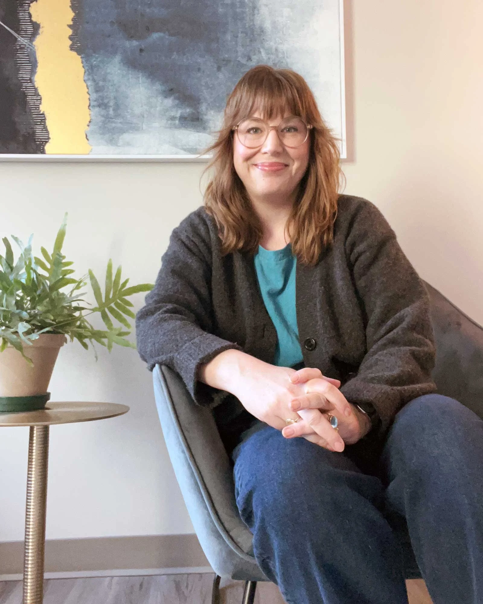 Crystal Tracy with shoulder-length wavy hair and glasses sits at a table, smiling, with a potted plant on the left and a colorful abstract painting on the wall behind her.
