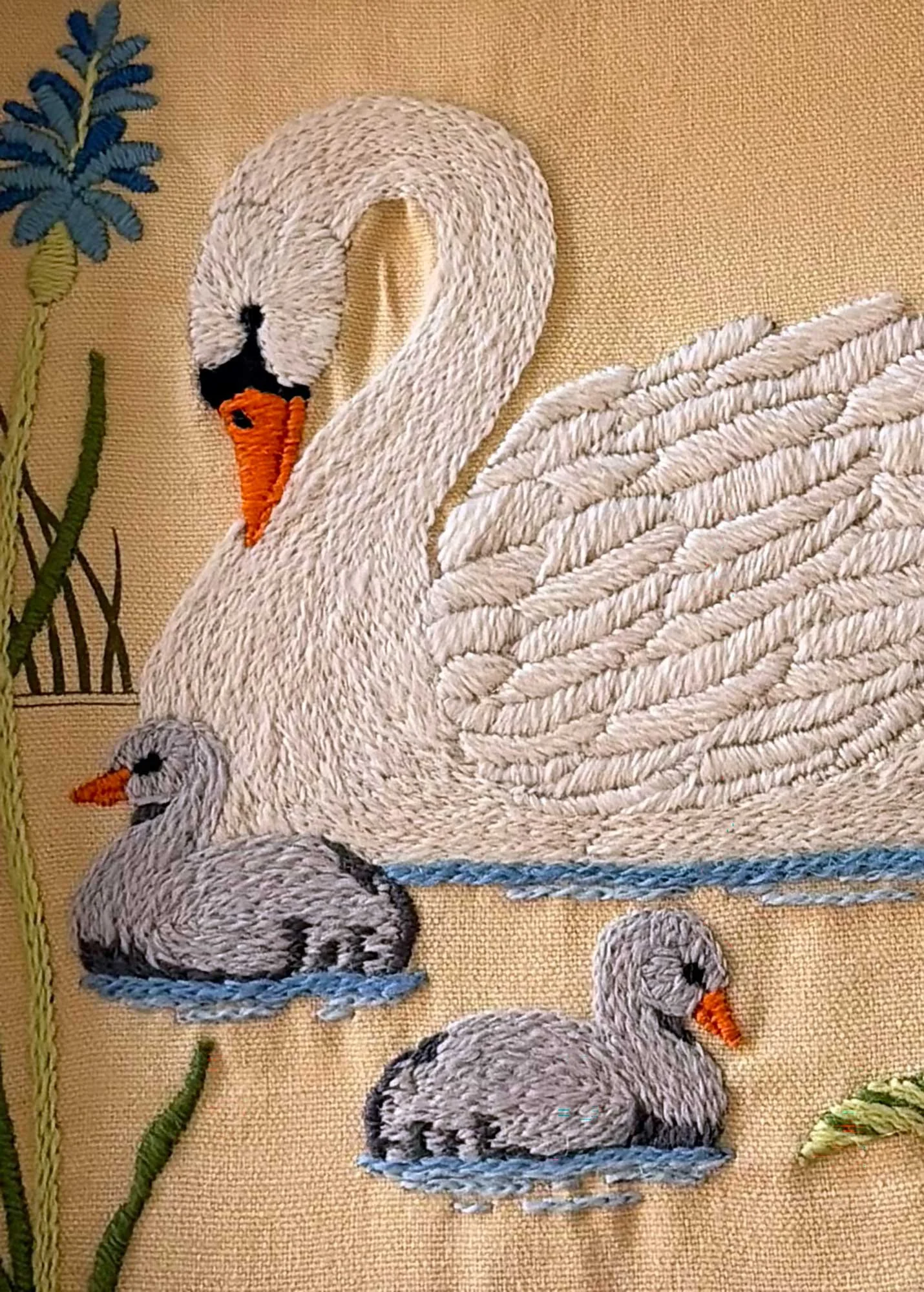 Embroidery of a swan and babies on a beige fabric background, with embroidered flowers and reeds.