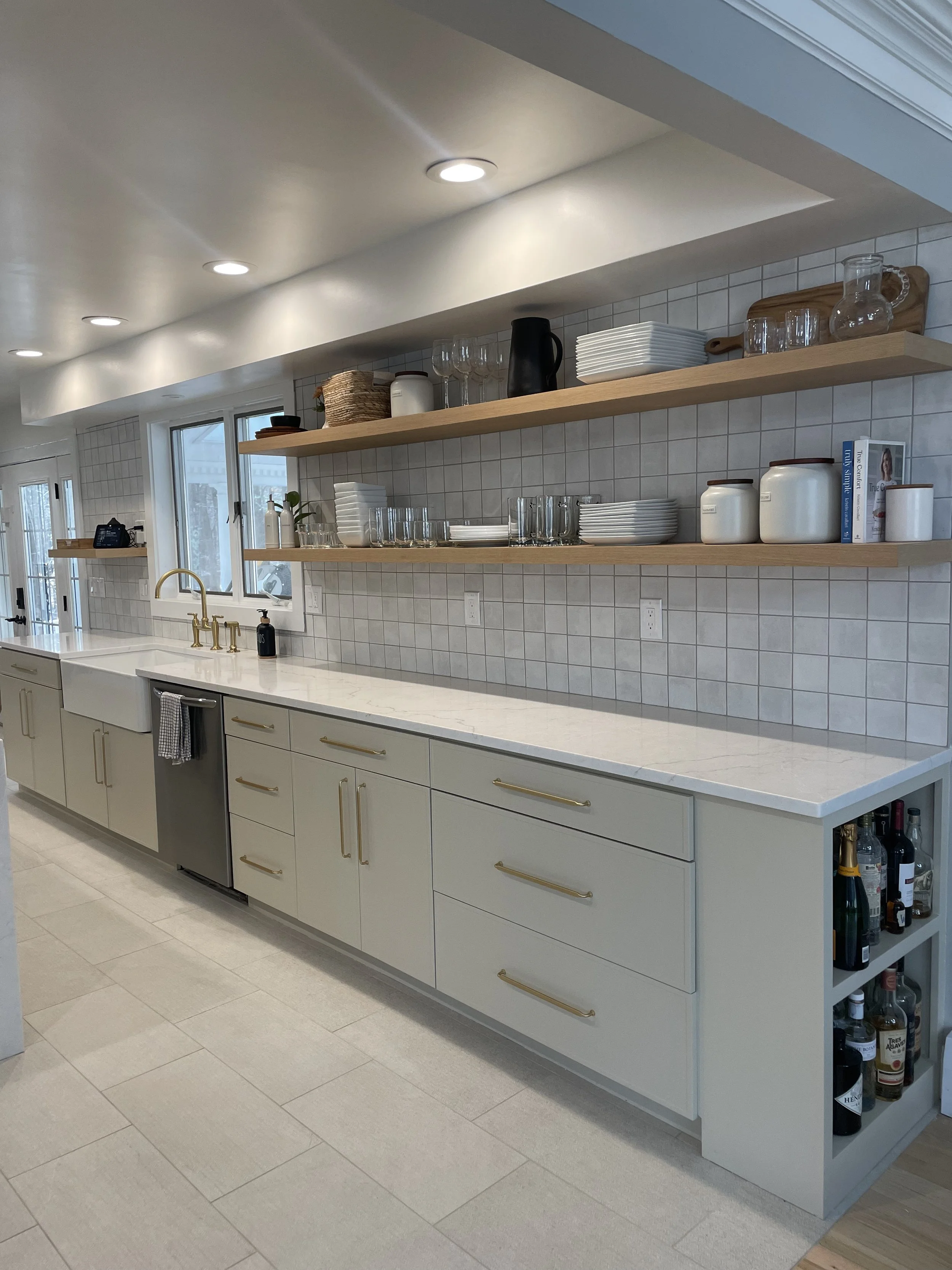 Modern kitchen with white cabinetry, open wooden shelves with dishes and glassware, a large window, beige tile backsplash, under-cabinet and recessed ceiling lighting, and a built-in wine storage area.