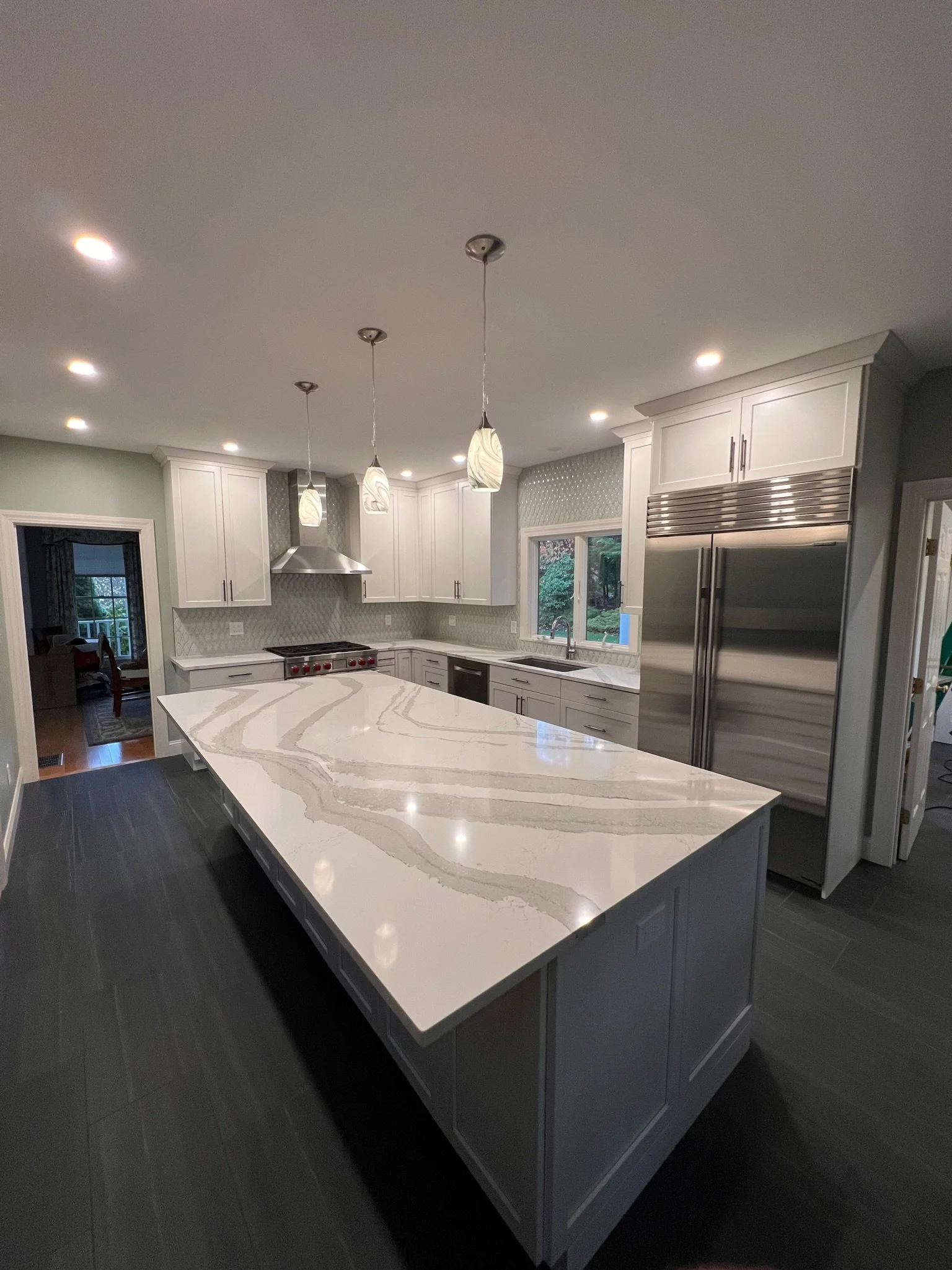 Modern kitchen with white cabinets, a large marble countertop island, stainless steel appliances including a double-door refrigerator, pendant lights above the island, and a window overlooking a garden.