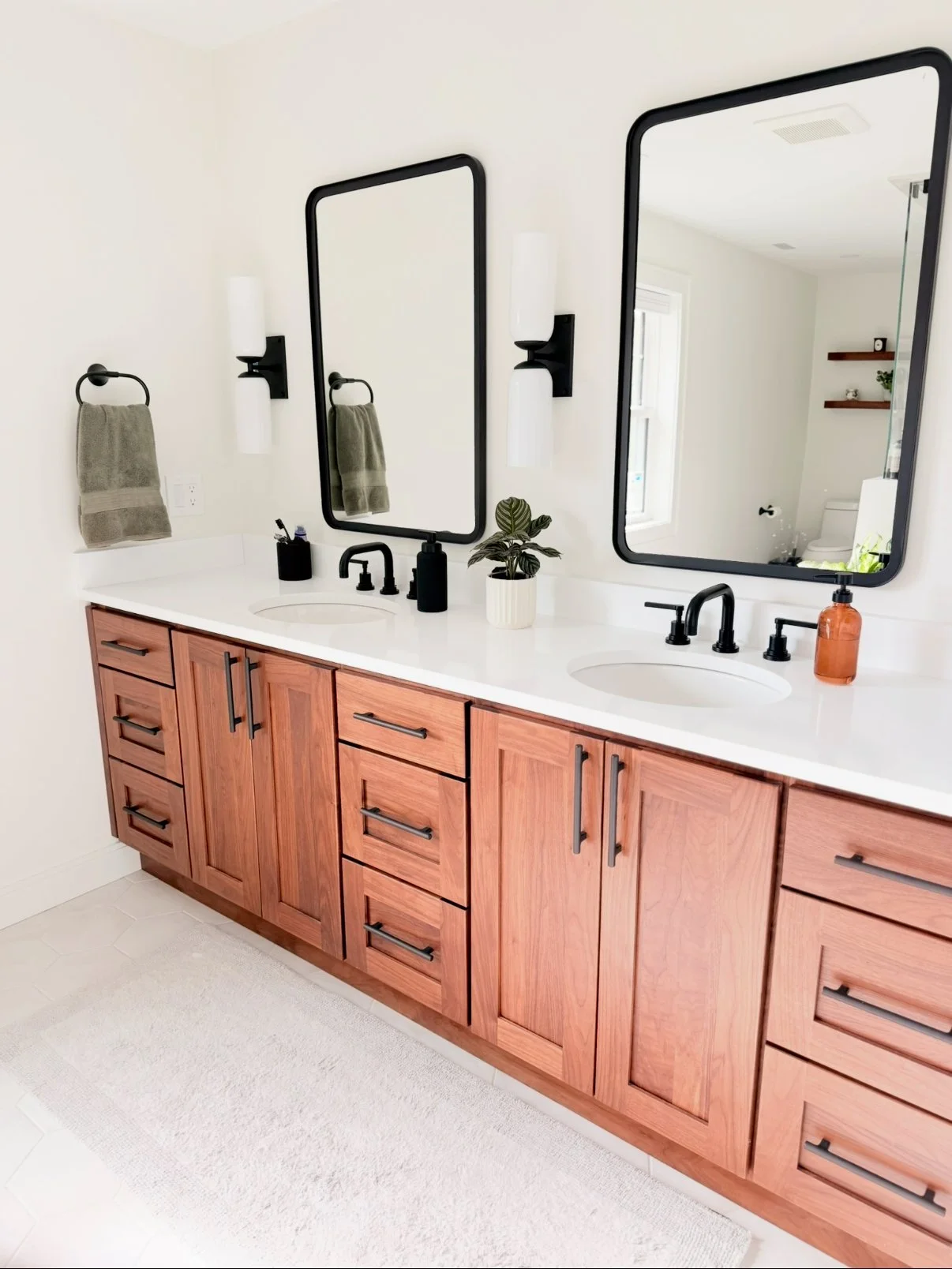 Bathroom vanity with two sinks, black fixtures, wooden cabinets, three mirrors, wall-mounted light fixtures, a potted plant, and soap dispenser.