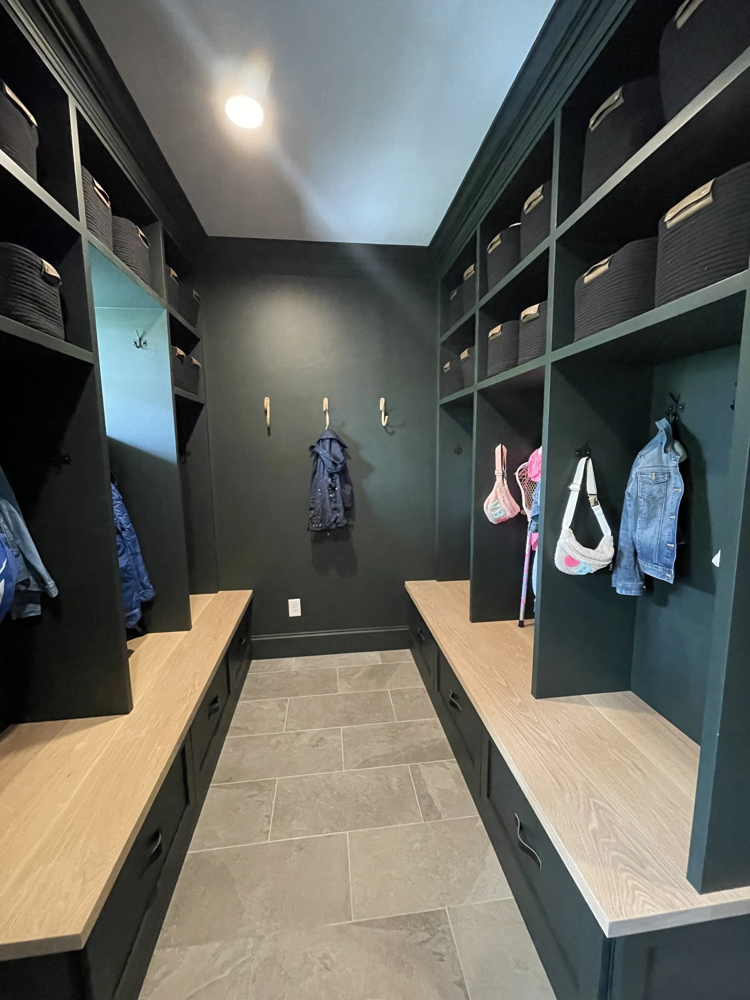 Mudroom with dark green built-in shelves and cubbies, with hooks holding jackets and bags, wooden benches, and gray tile flooring.