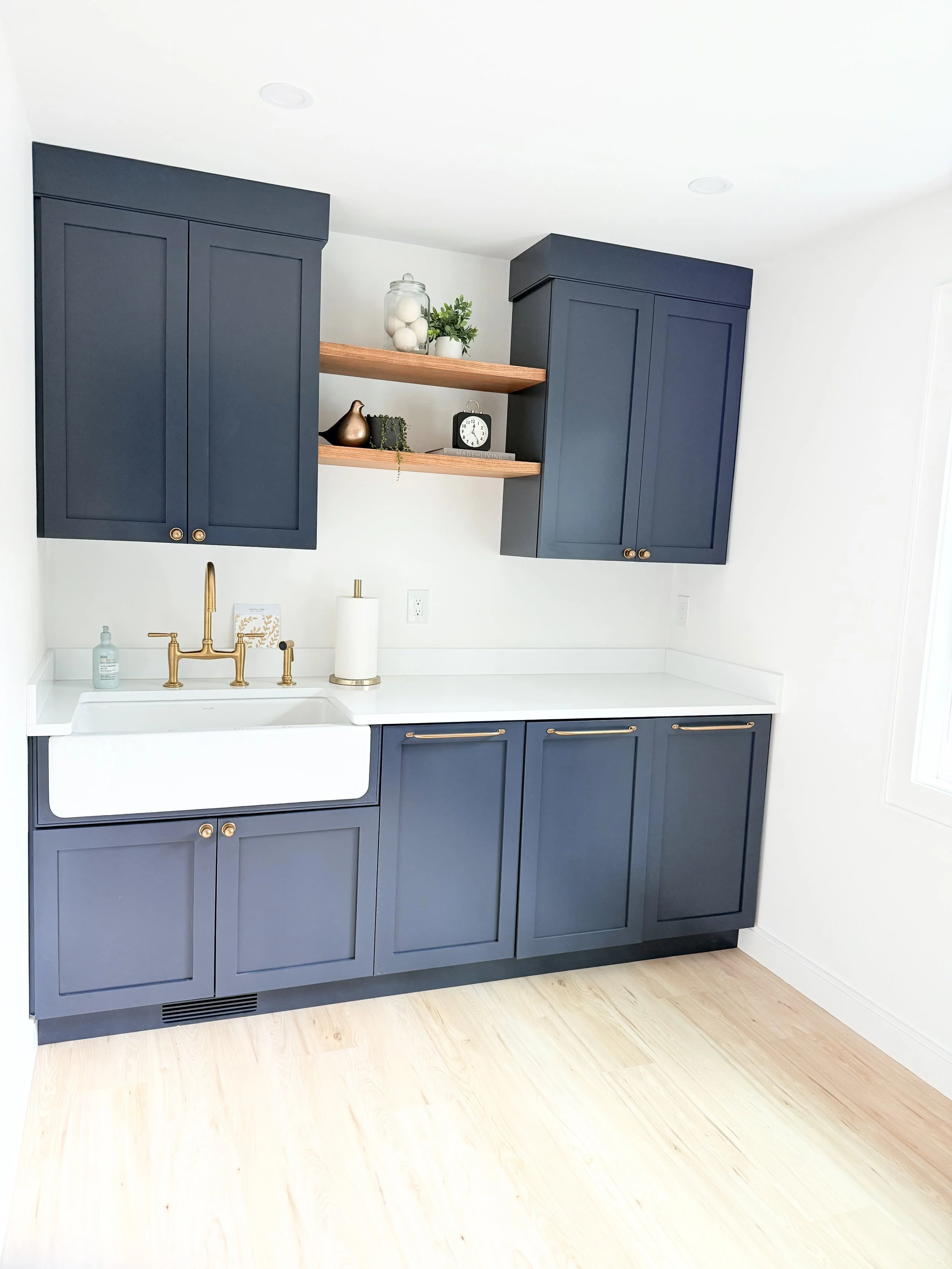 Laundry room with navy blue cabinets, a white sink with a gold faucet, a wooden shelf with decorative items, and a light-colored wooden floor.