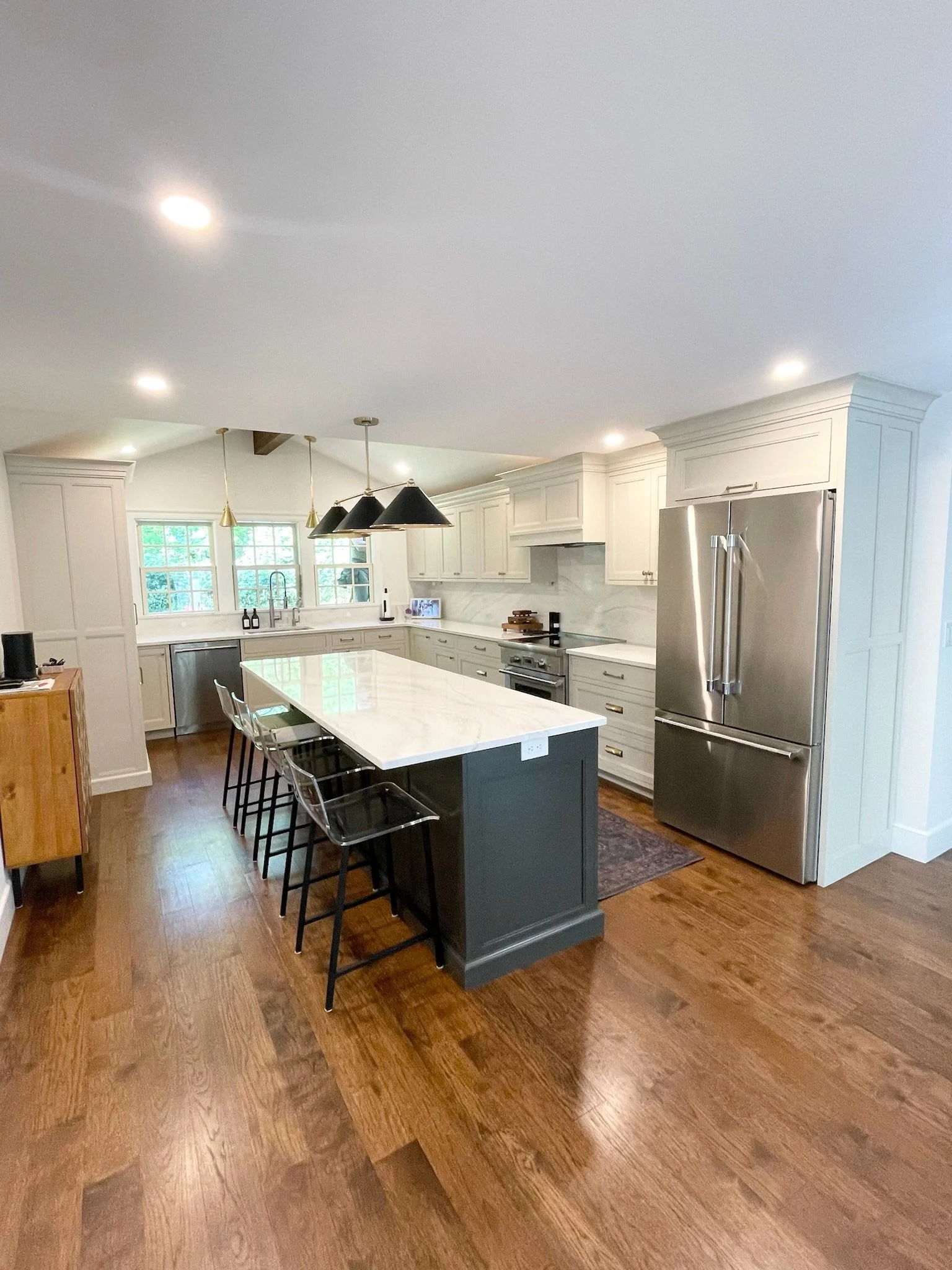 Modern kitchen with white cabinets, stainless steel refrigerator, kitchen island with marble countertop, black bar stools, hardwood floors, and pendant lights.