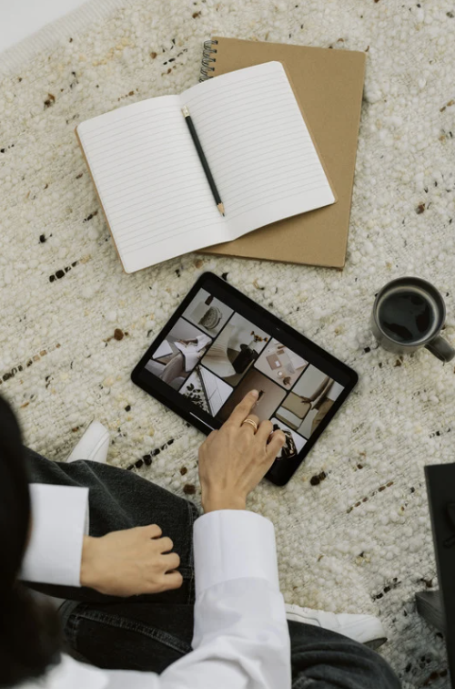 Person sitting on a carpeted floor, using a tablet to browse photos, with a cup of coffee, an open notebook with a pencil, and a closed notebook nearby.