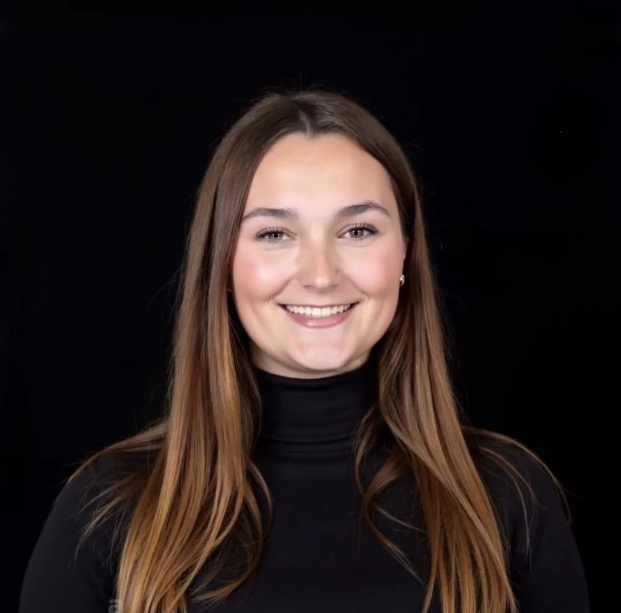 A young woman with long brown hair smiling, wearing a black turtleneck, against a plain black background.