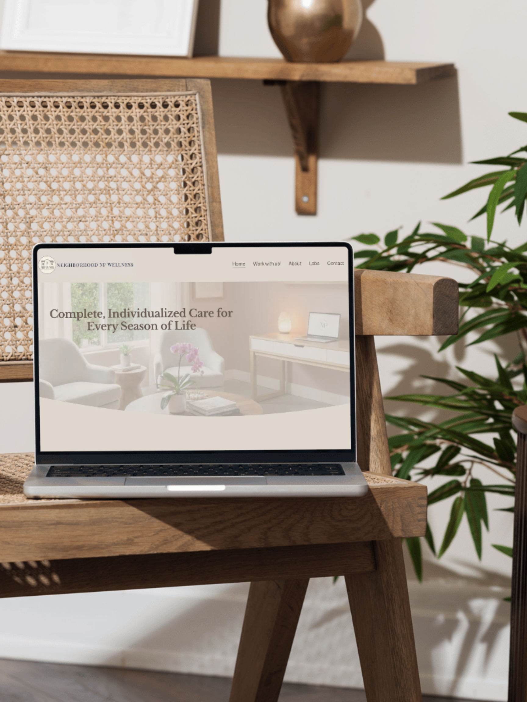 Laptop on a wooden table displaying a wellness website with a living room background, surrounded by a home interior with a wicker and wood wall shelf and green plant.