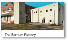 Exterior view of The Barnum Factory, a white industrial building with small windows and a concrete structure, with a parking lot in front.