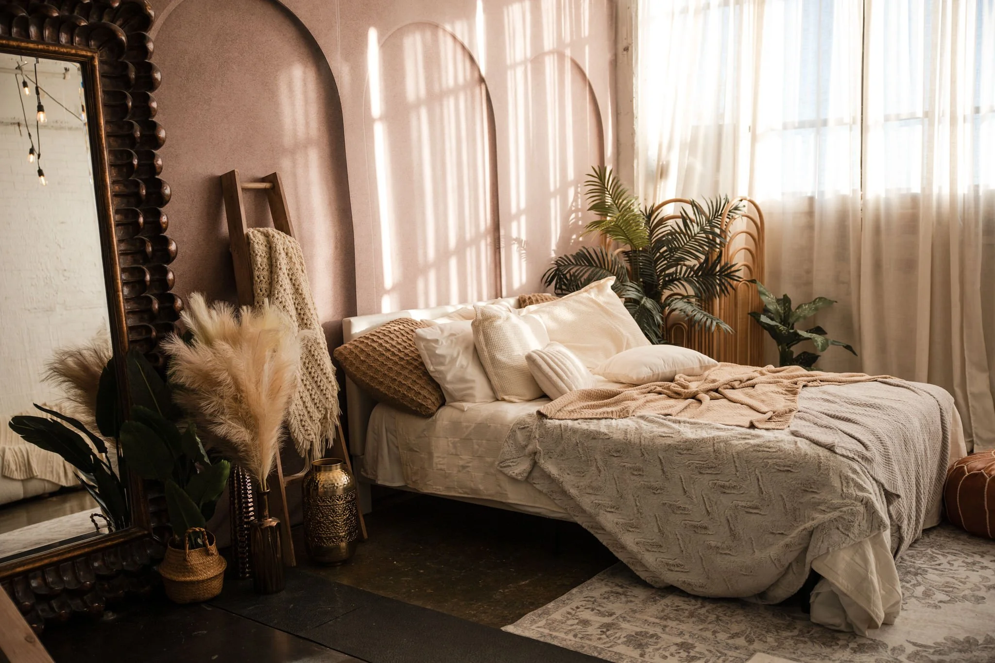 A cozy bedroom with sunlight streaming through large sheer curtains. The bed is decorated with various textured pillows and a knit blanket. There are indoor plants, a large ornate mirror, a wooden ladder with a throw blanket, and decorative vases on the floor.