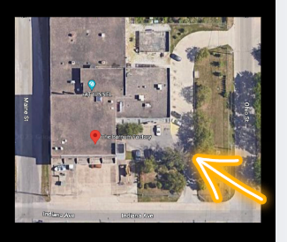 Aerial view of a building with surrounding streets, trees, and parking area, marked with a red location pin and an orange arrow pointing to a sidewalk.