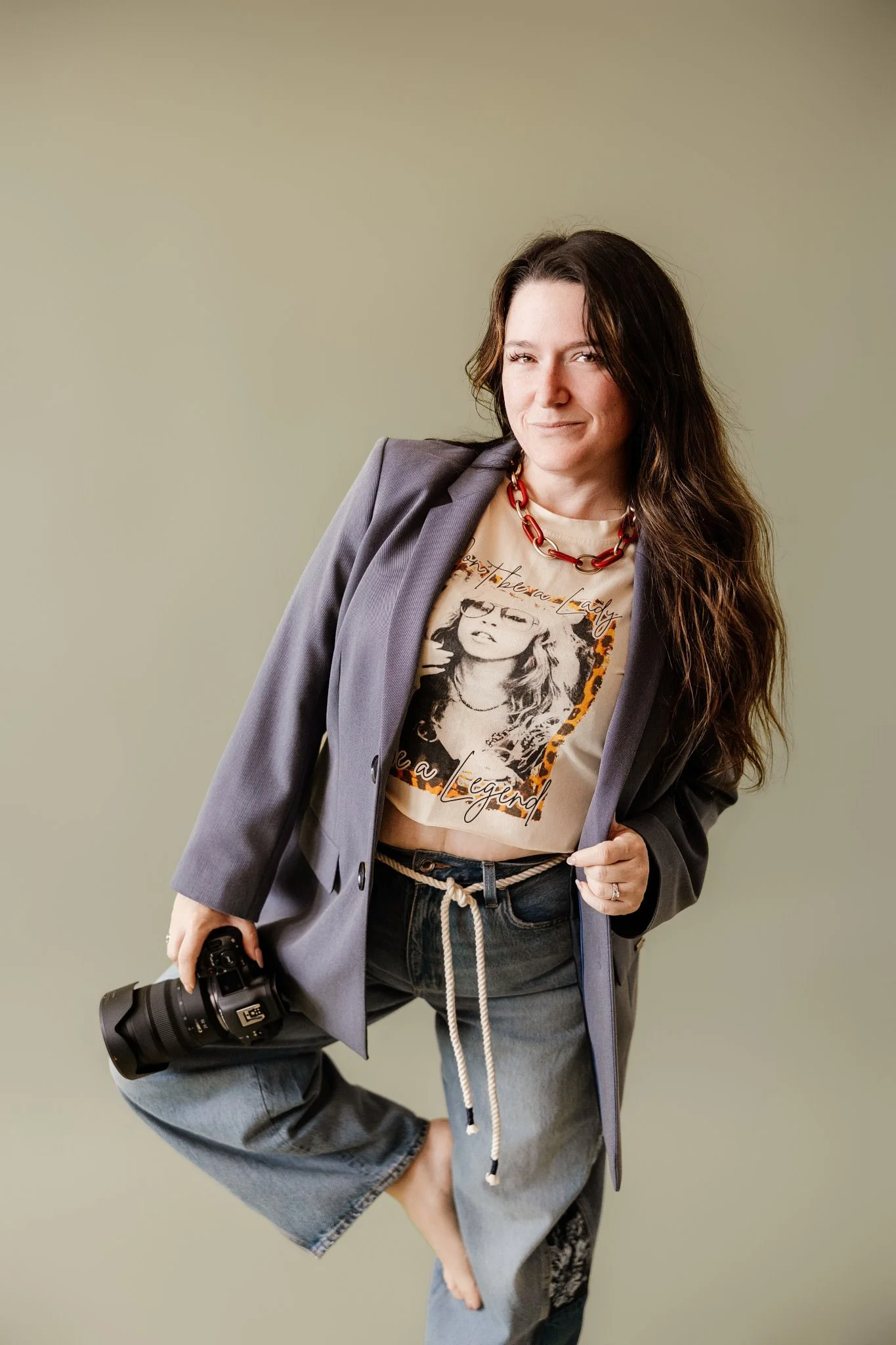 A woman with long dark hair wearing a gray blazer, a graphic t-shirt, and jeans, holding a professional camera in her right hand, standing against a plain, light olive background.