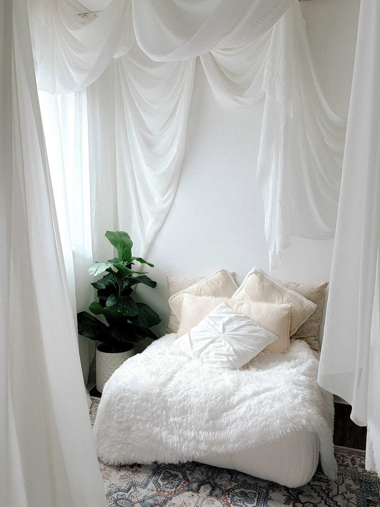 A cozy, white, canopy escape with a plush fluffy rug, multiple cream-colored pillows, a potted green plant, and sheer white curtains draped over a bed or seating area.