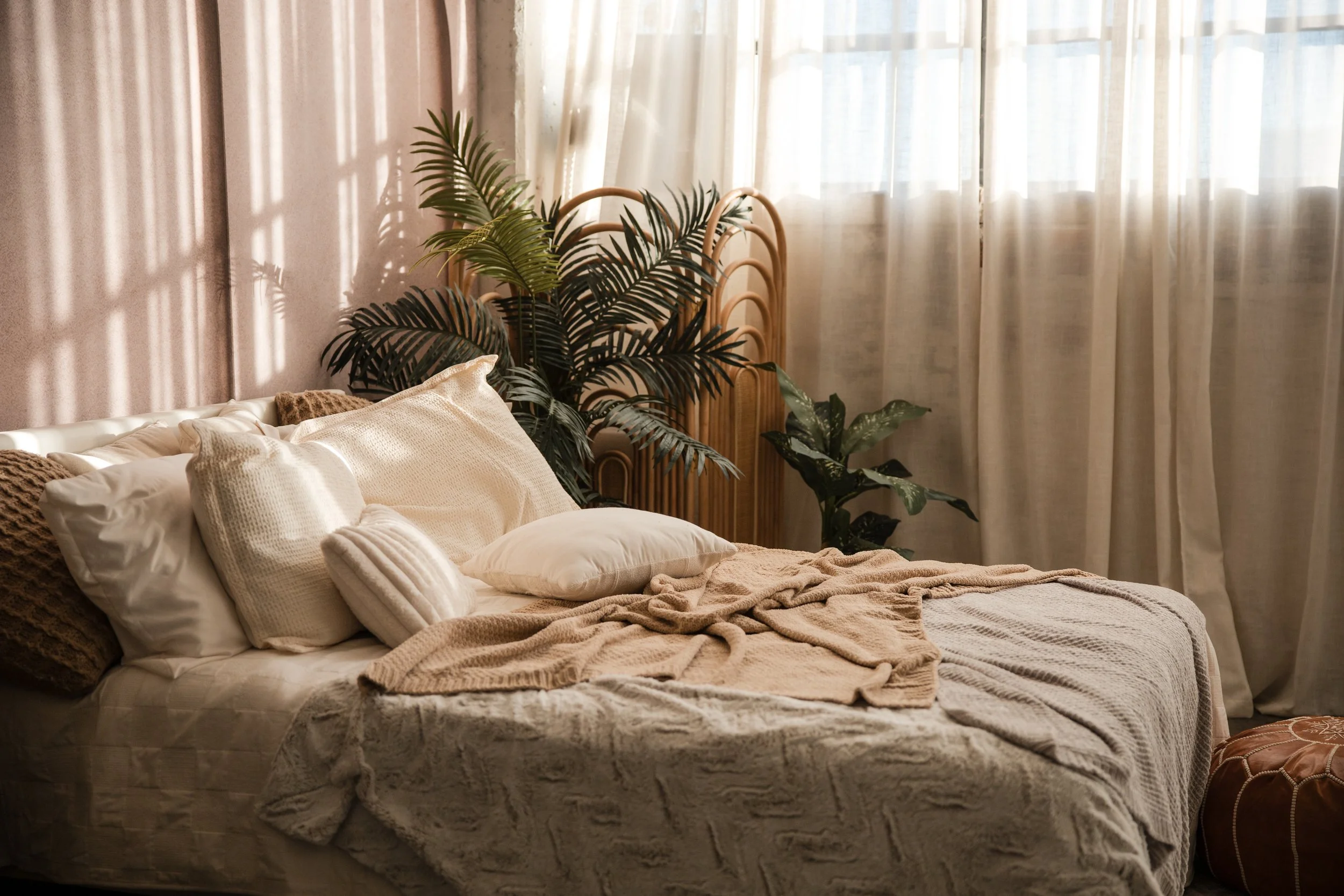 A cozy bedroom with a bed covered in beige and white blankets and pillows. There are large green plants next to a rattan headboard, with sheer curtains letting in soft natural light.
