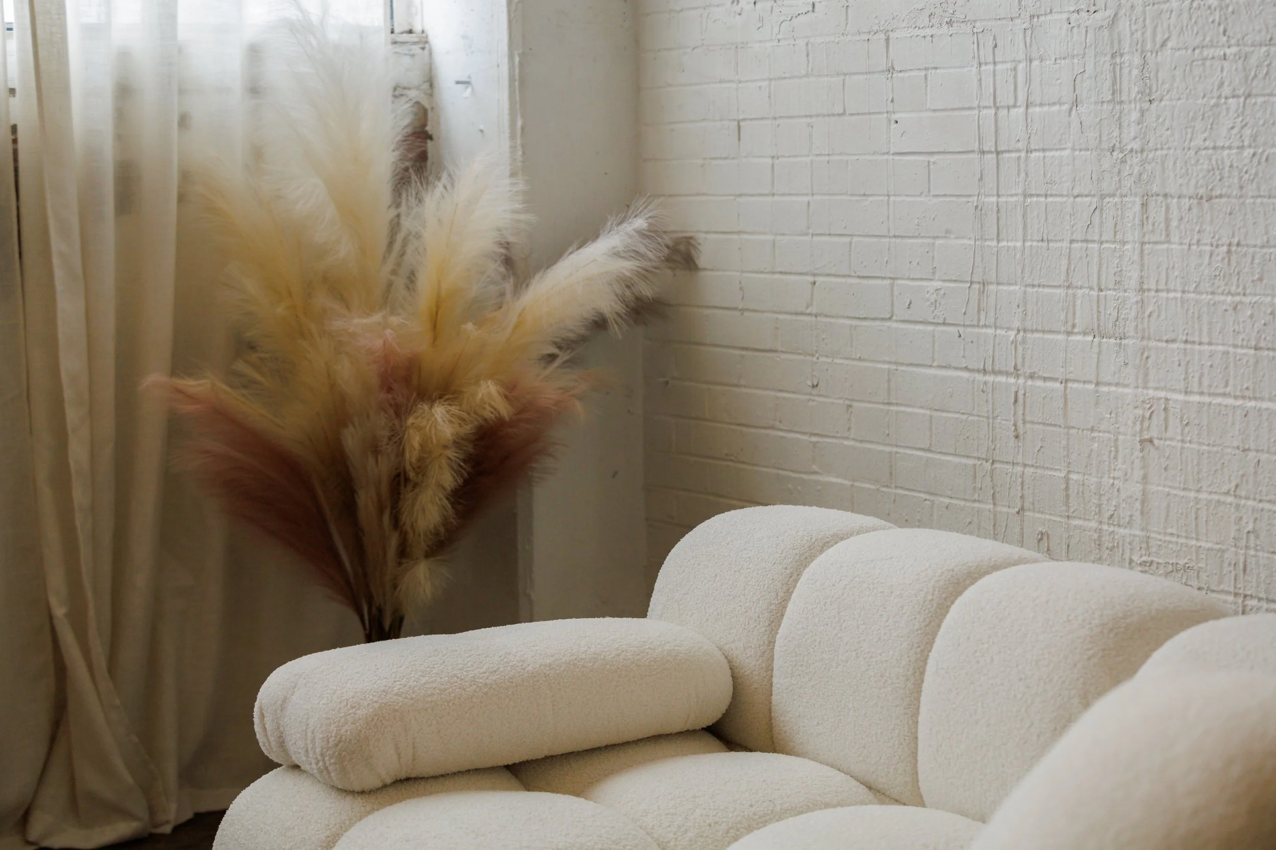 A fluffy beige and brown feather duster leaning against a white brick wall next to a beige upholstered chair with rolled arms.