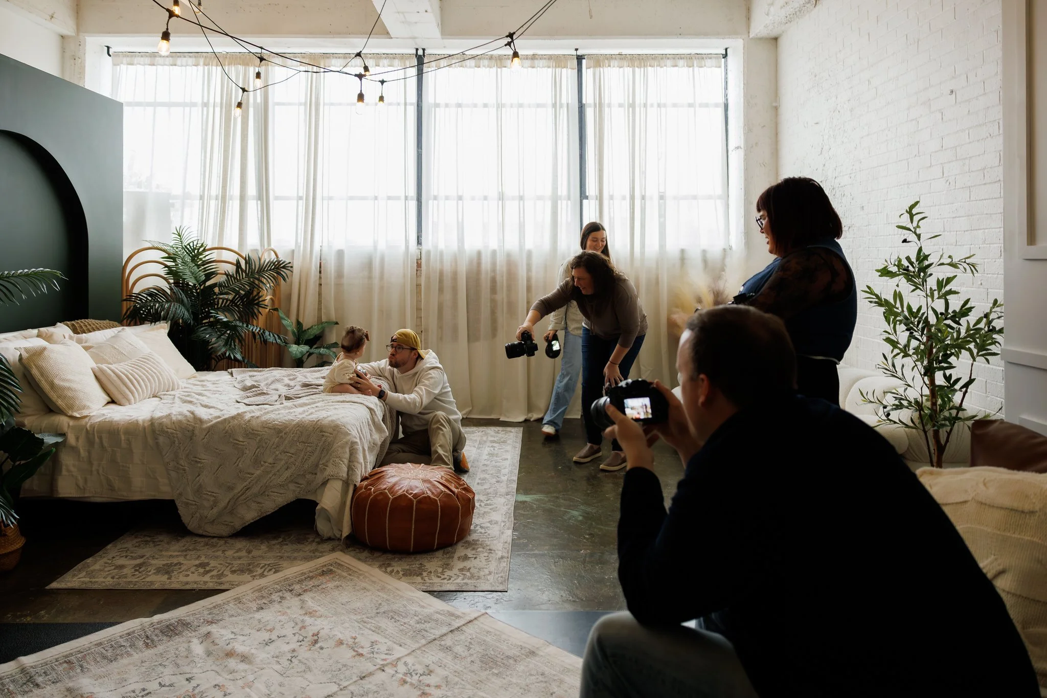 A group of people in a well-lit room, possibly a photo shoot, with a child sitting on a bed and adults around taking pictures. The room has large windows with sheer curtains, plants, and a hanging light fixture.