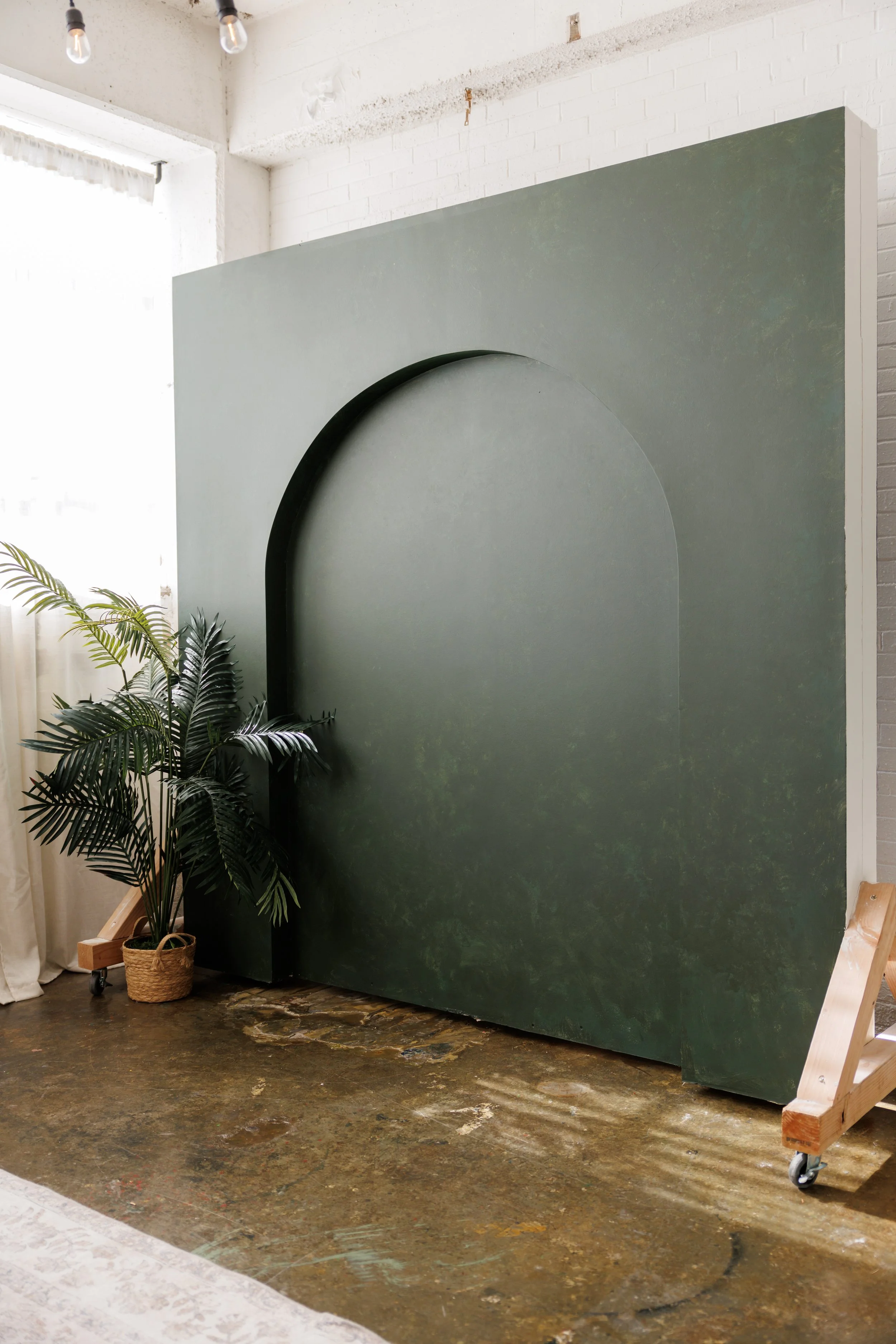 A green wall with an arch design, placed indoors near a white brick wall and a window with natural light. There are plants in baskets on the floor and a wooden bench on wheels nearby.