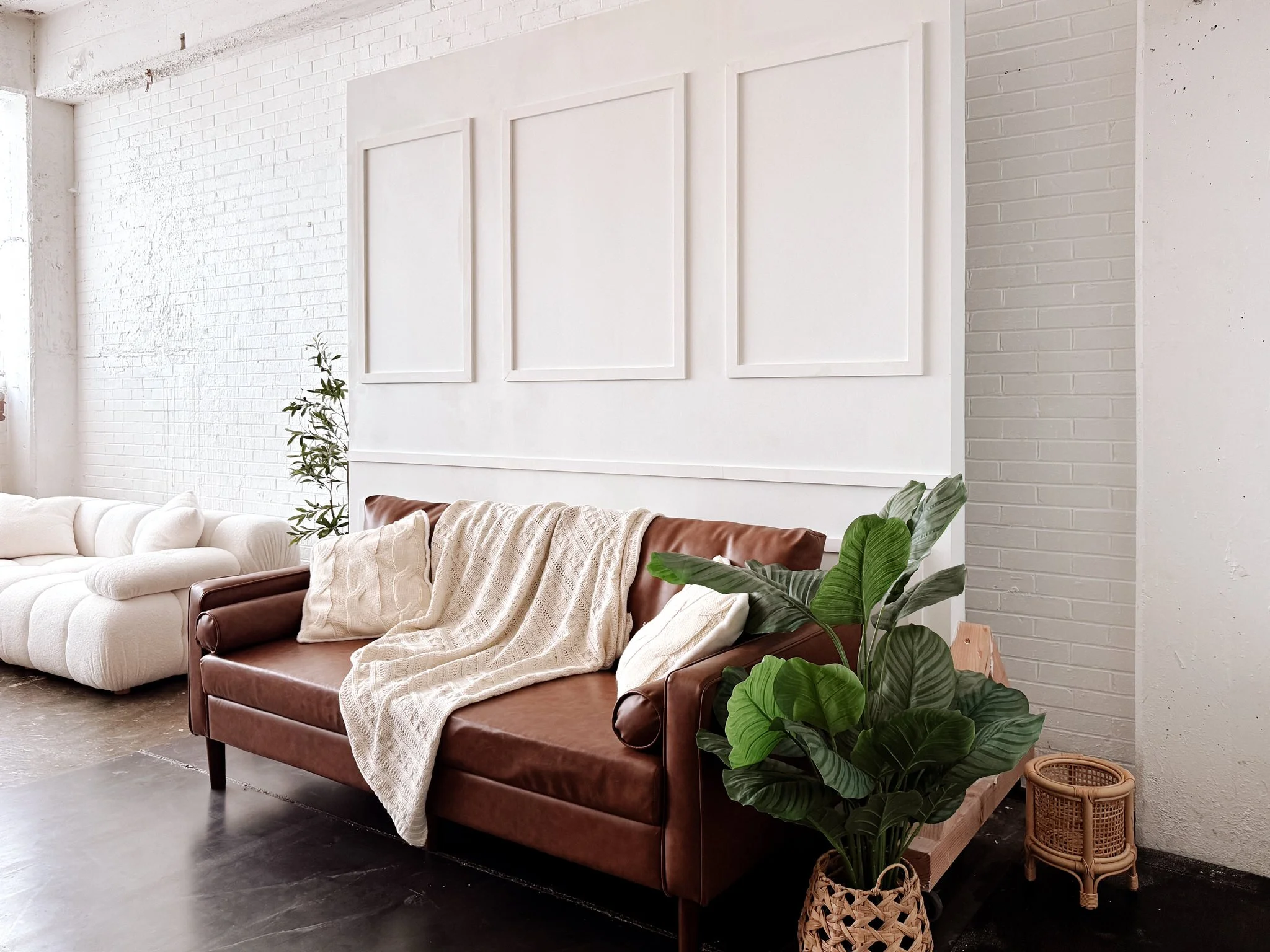 Interior of a modern living room with a white brick wall, a white sofa, a brown leather sofa with a blanket, green plants, a small woven basket, and picture frames on the wall.