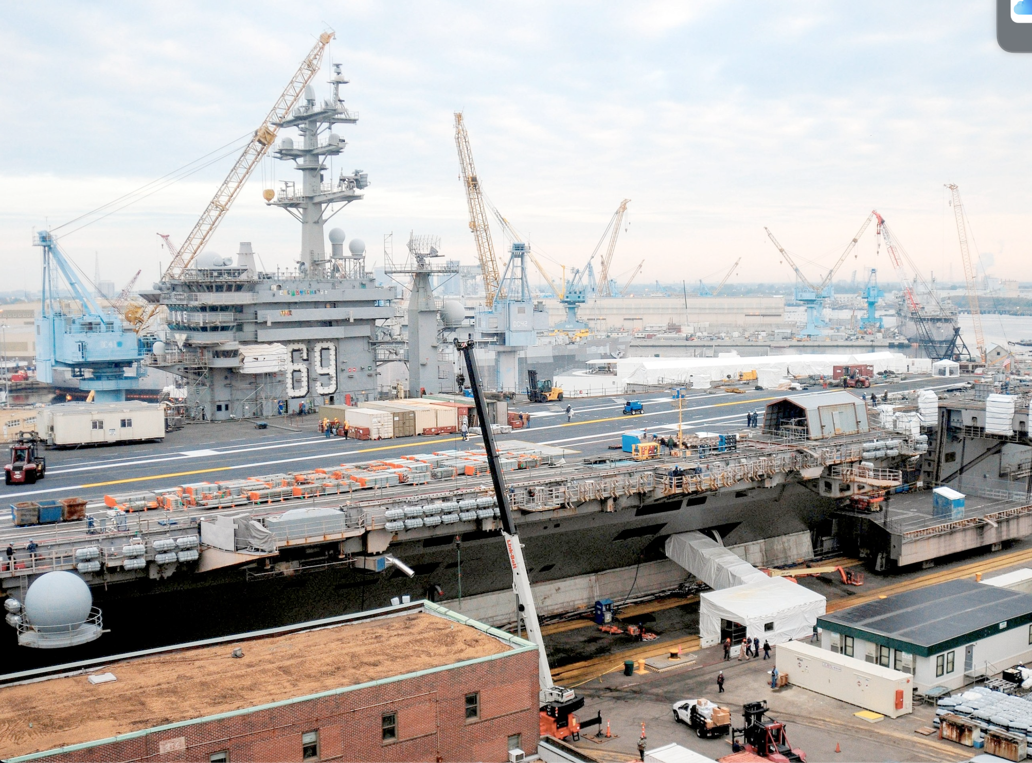 View of an aircraft carrier under construction or maintenance with cranes and workers on deck.