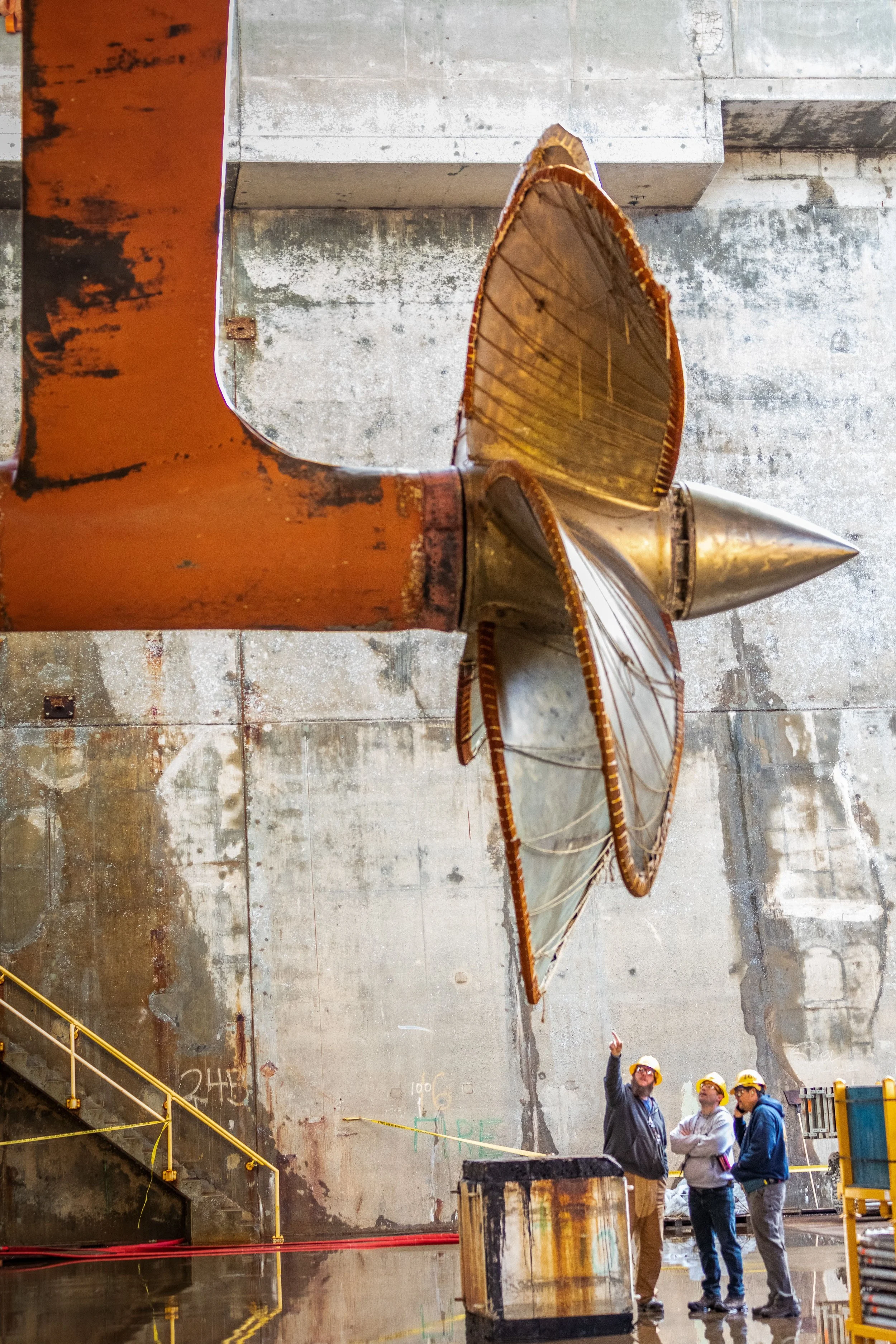 A group of people wearing safety helmets in an industrial setting, standing near a large rusty metallic structure that resembles an aircraft component or industrial equipment. The background features a large concrete wall and a staircase on the left. One person is pointing upward, likely explaining or inspecting the equipment.