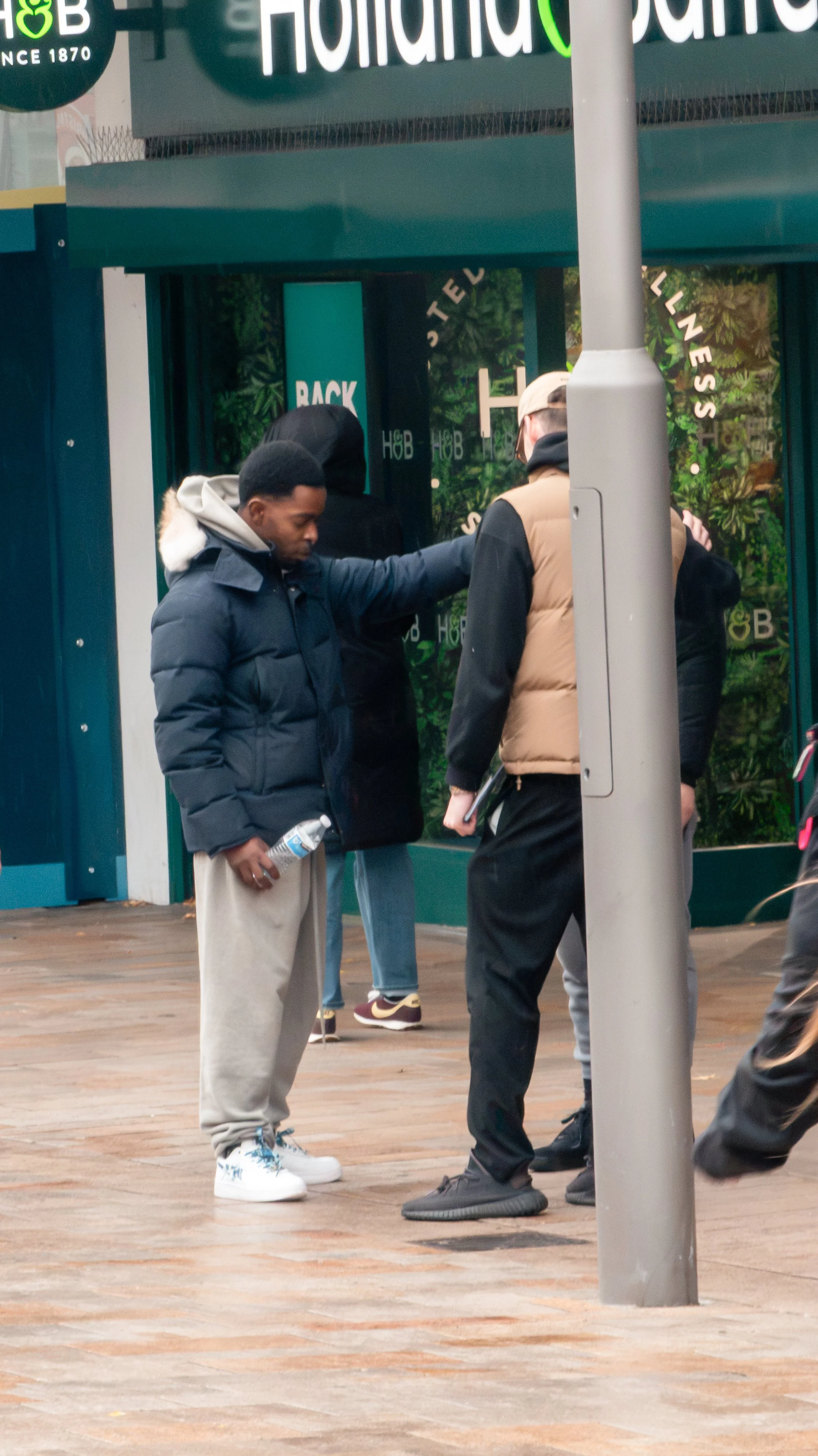 People in winter clothing walking outside a storefront with green signage and white columns.