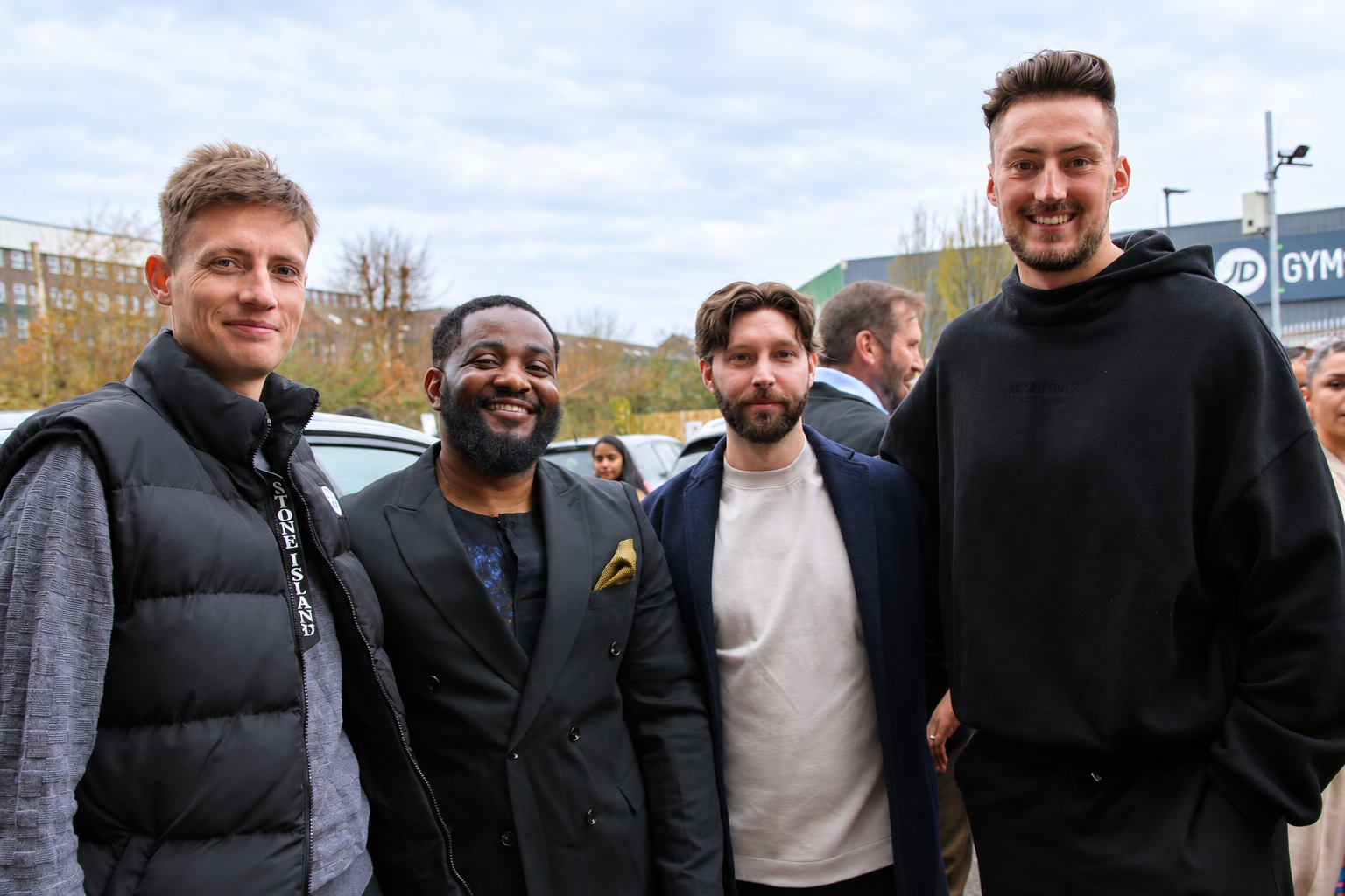 Four men pose together outdoors in front of a gym and parked cars.