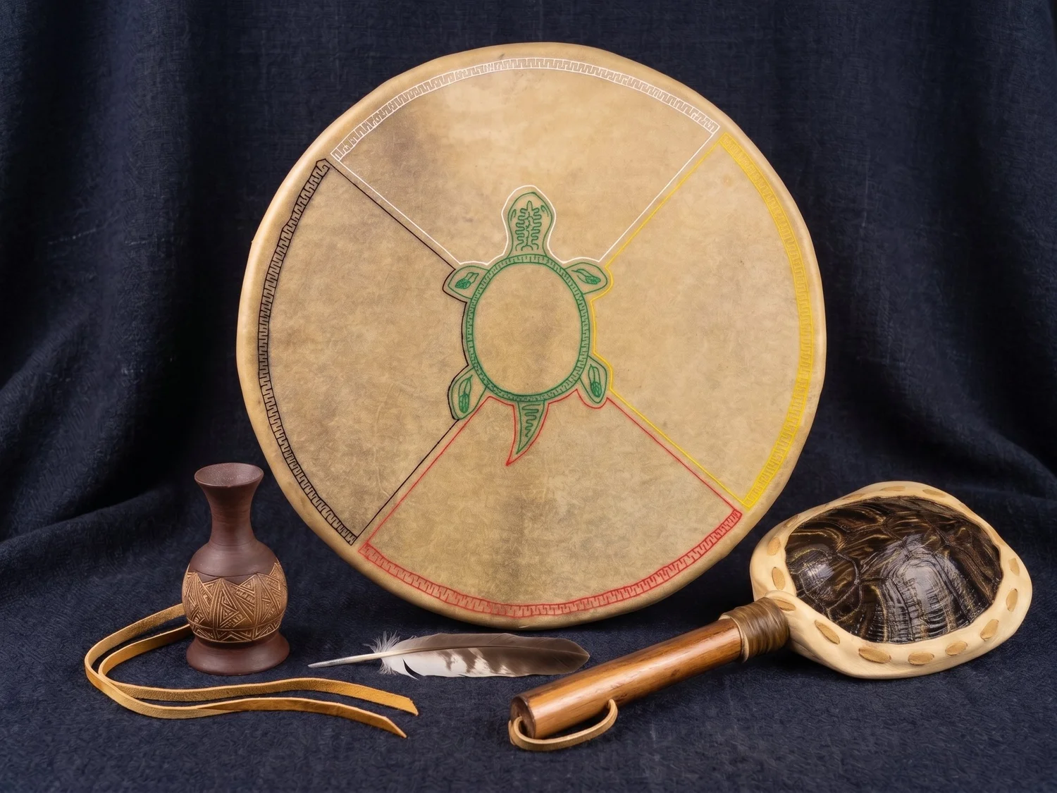 A hand drum with a green turtle at its centre and four coloured sections in black, white, yellow and red, displayed with a turtle shell rattle, a feather and a small clay vase on dark blue fabric