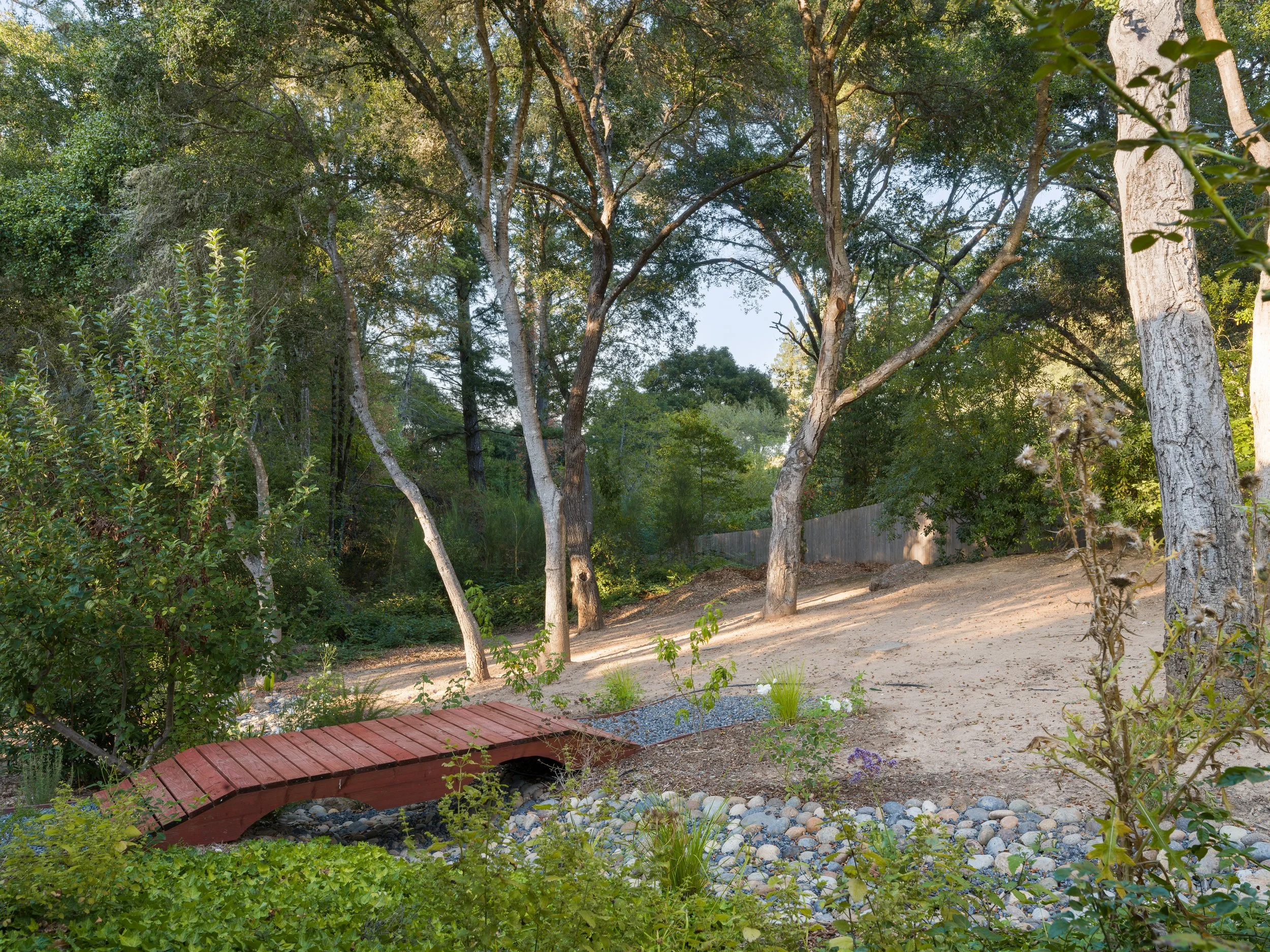 A backyard with a small wooden bridge over a dry creek bed, surrounded by various green plants and trees, with a dirt path and a wooden fence in the background.