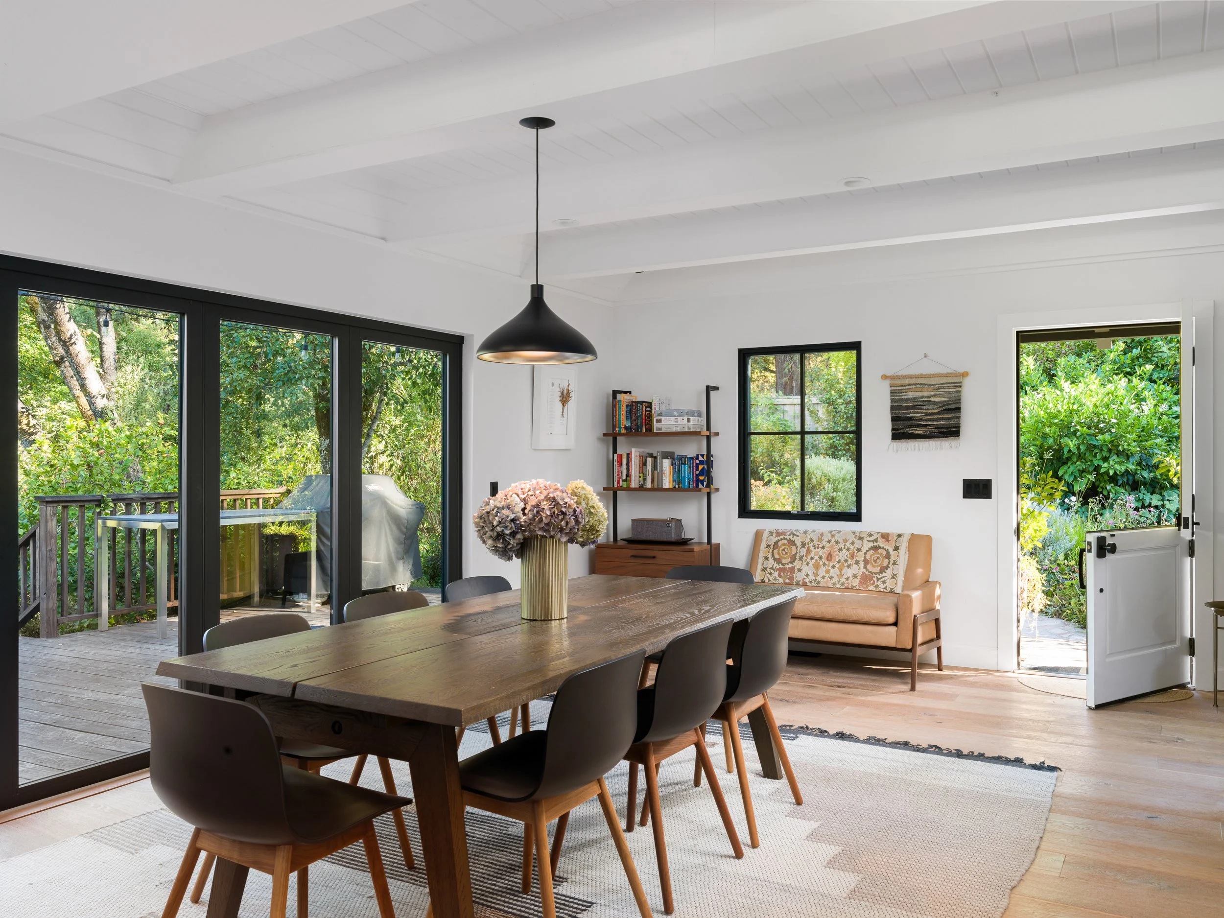 Interior view of a dining area with a wooden table, black chairs, a flower vase, bookshelf, armchair, wall art, and large windows with outdoor greenery.