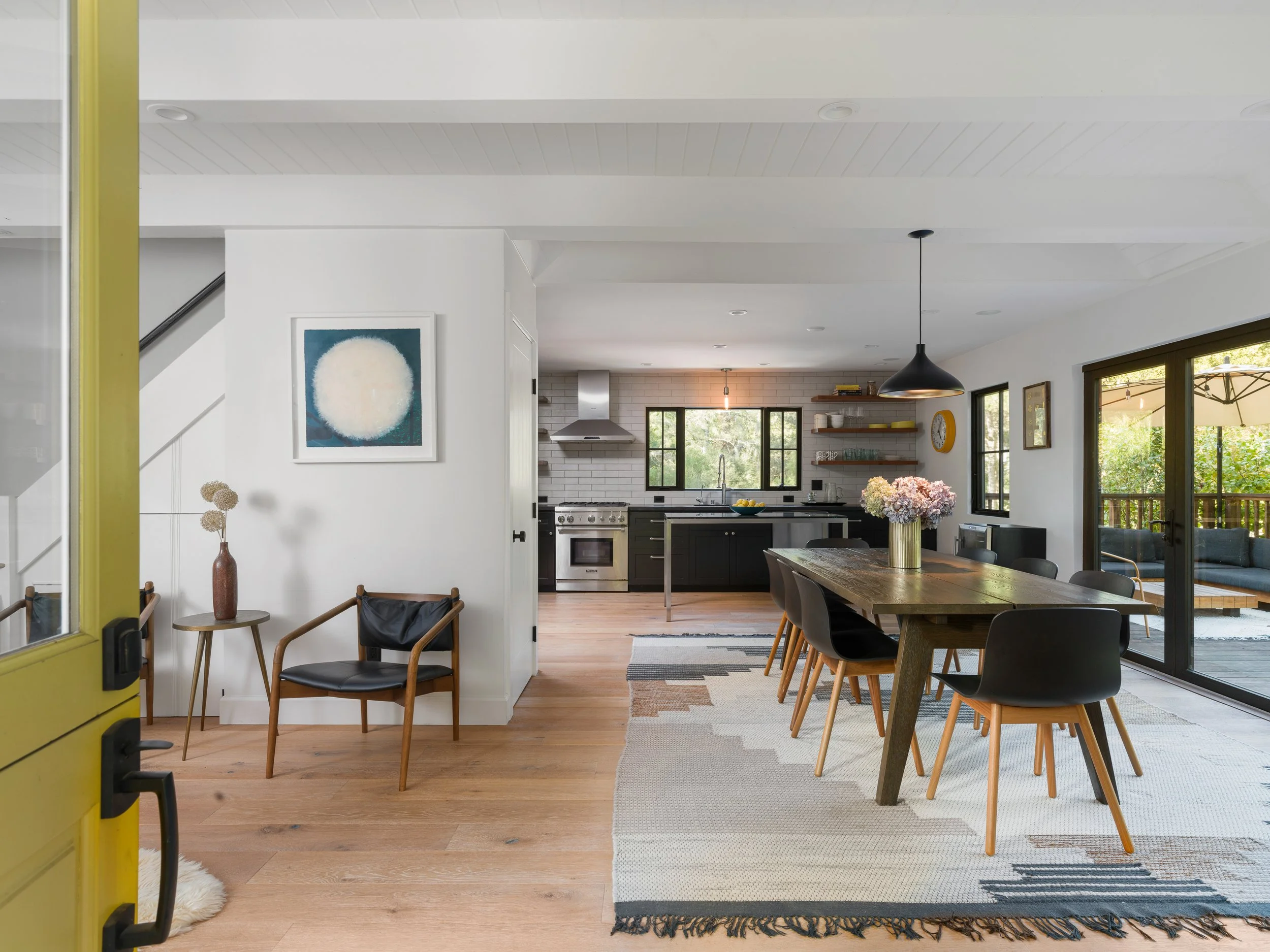 Modern open-plan kitchen and dining area with white walls, black cabinets, wooden dining table, and large windows showing greenery outside.