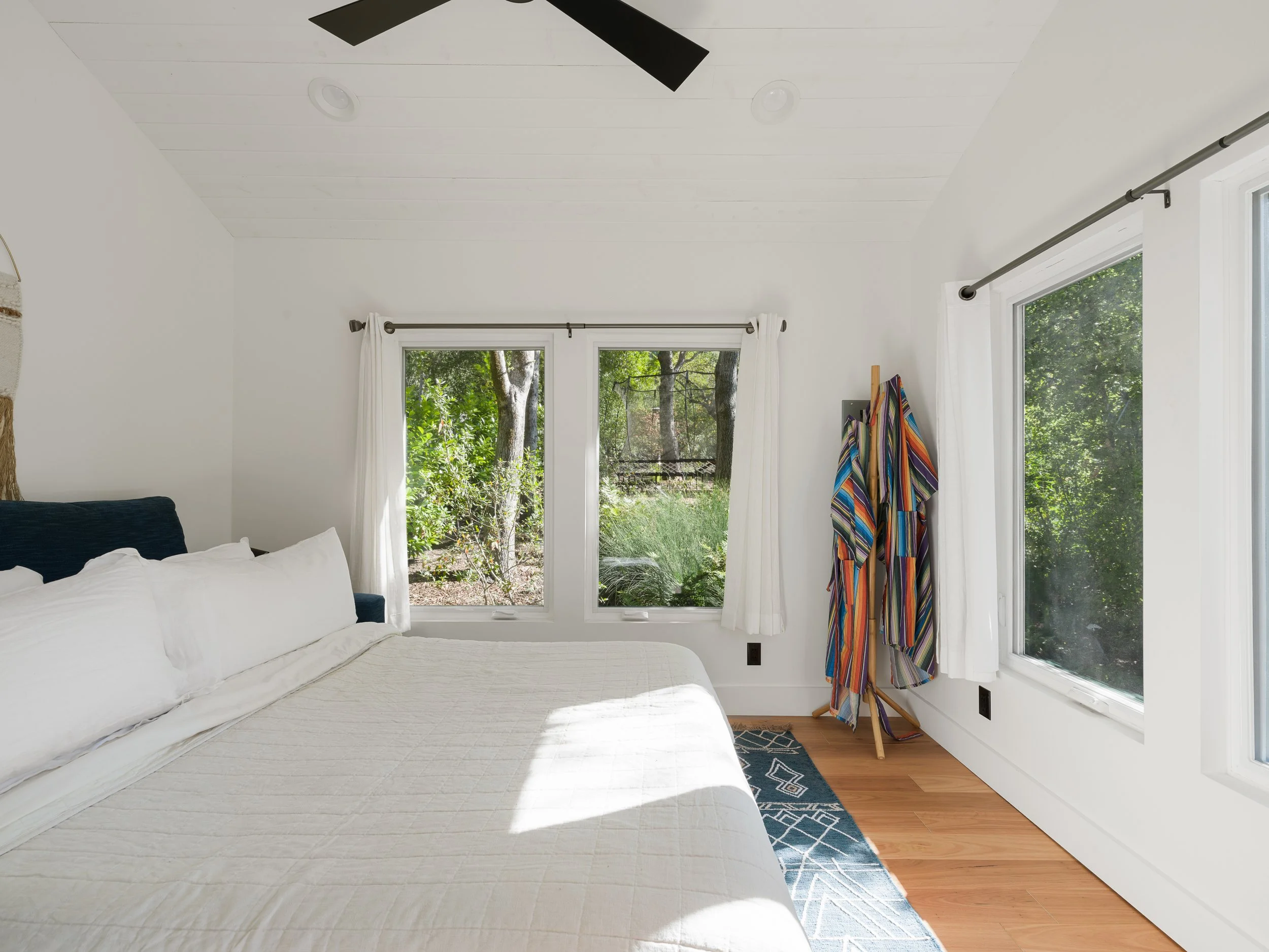 A bedroom with white walls, a white ceiling, and hardwood floors. There are two windows with white curtains, showing a view of green trees outside. Part of a bed with white bedding and a navy headboard is visible on the left. A striped colorful robe 