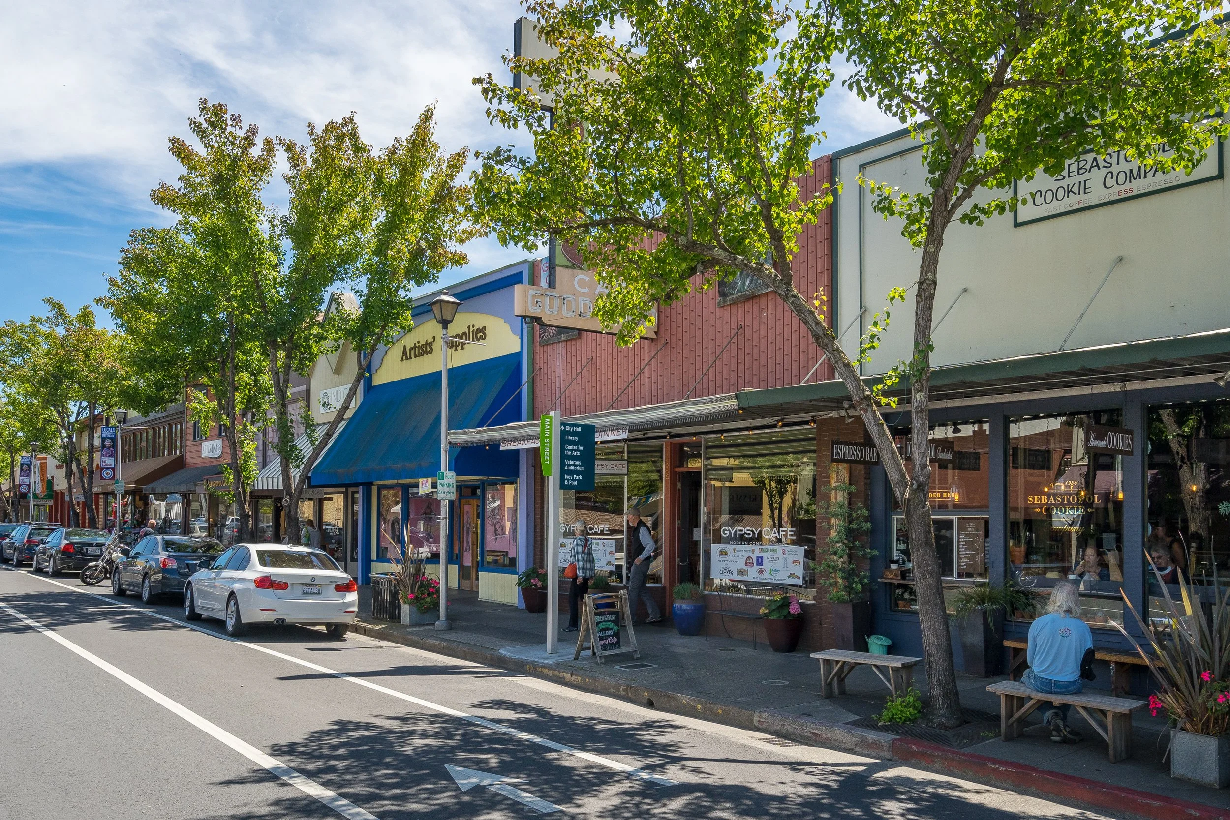 A street scene with parked cars in front of shopping and dining establishments. Green trees line the sidewalk, and pedestrians are walking or sitting outside. The buildings display colorful storefronts and signs for various businesses.
