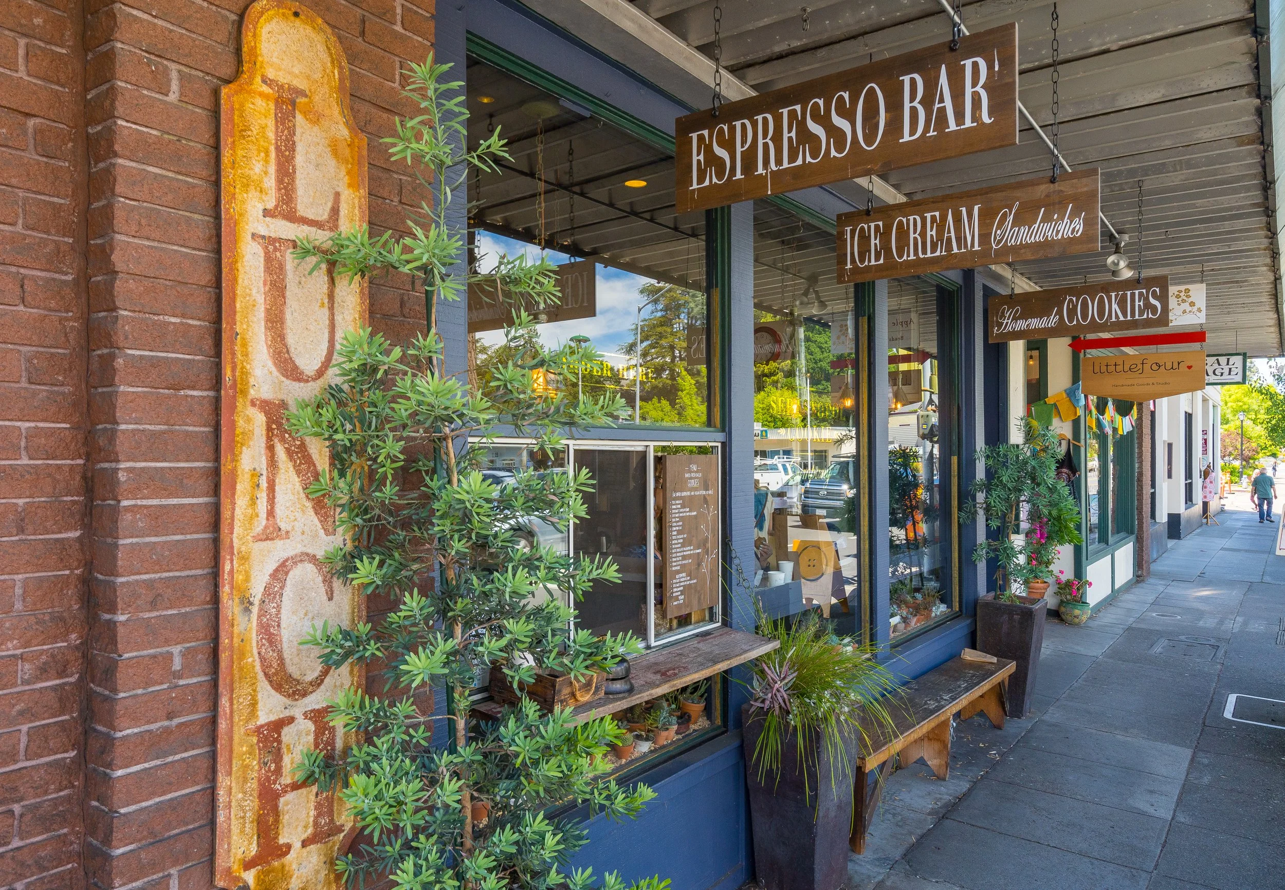 The image shows the exterior of a storefront with hanging signs that read 'Espresso Bar,' 'Ice Cream Sandwiches,' and 'Homemade Cookies.' There are plants and a decorative wooden bench outside, and a brick wall with a large, weathered metal sign on i
