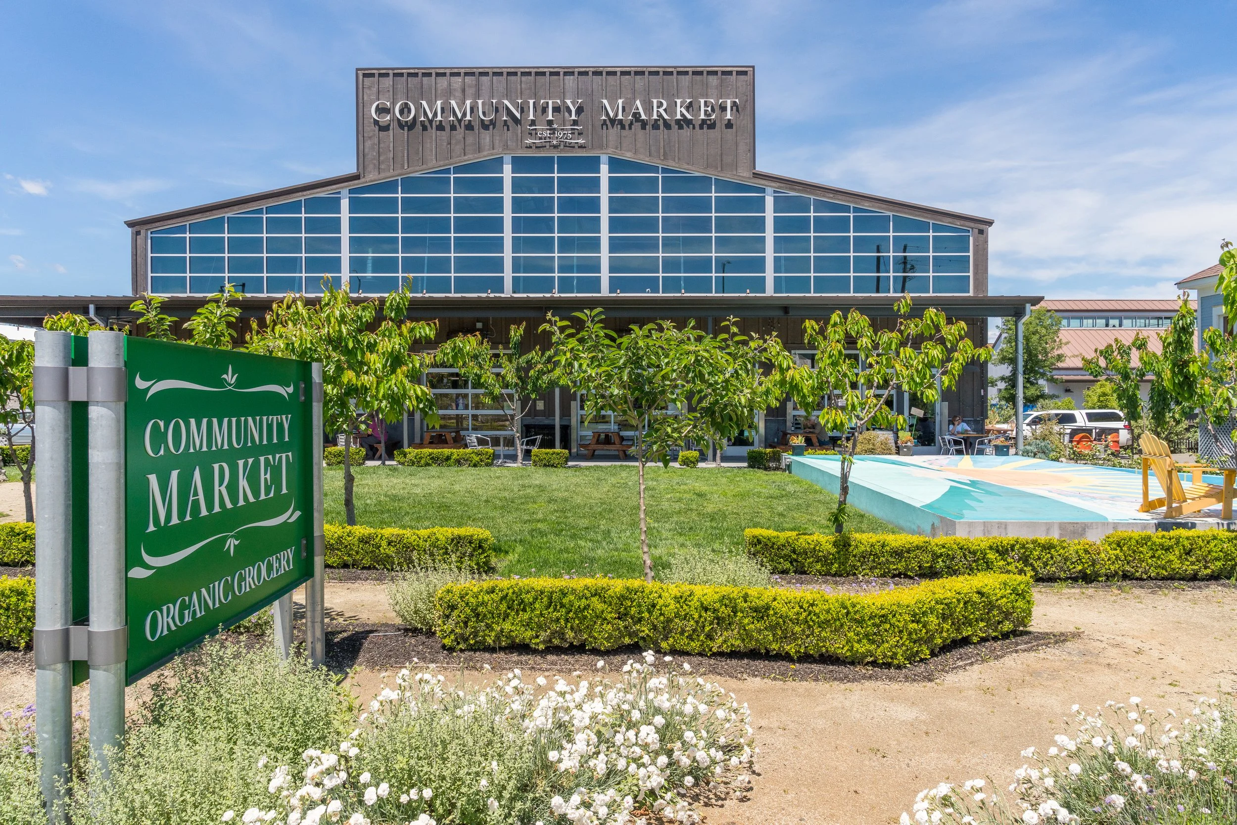 Exterior of a community market building with a sign in the foreground reading 'Community Market.' The market has large glass windows, a wooden upper facade, and surrounding greenery and seating areas.