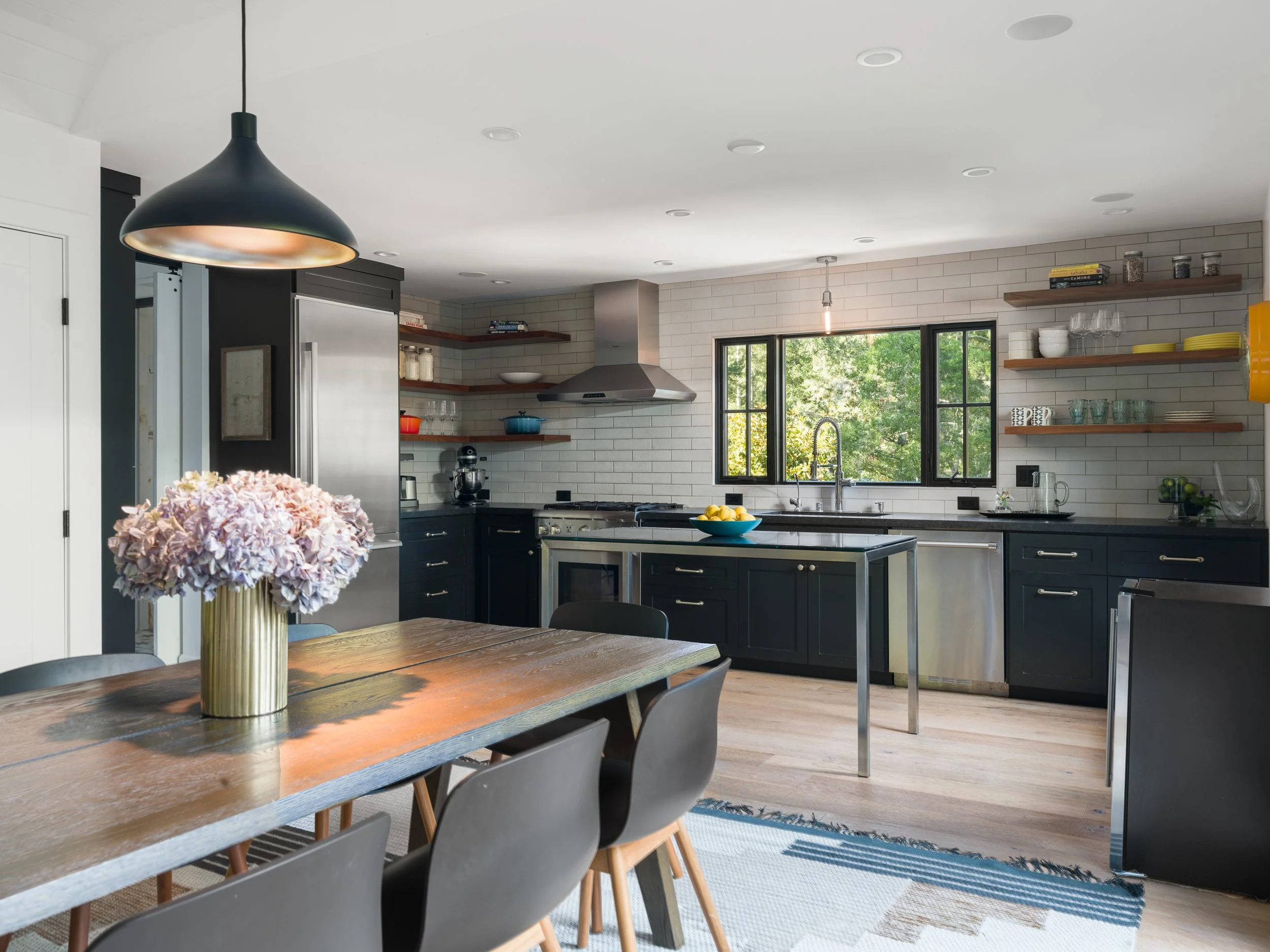 Modern kitchen with blue cabinetry, open wooden shelves, stainless steel appliances, and a dining table with a vase of pink hydrangeas.