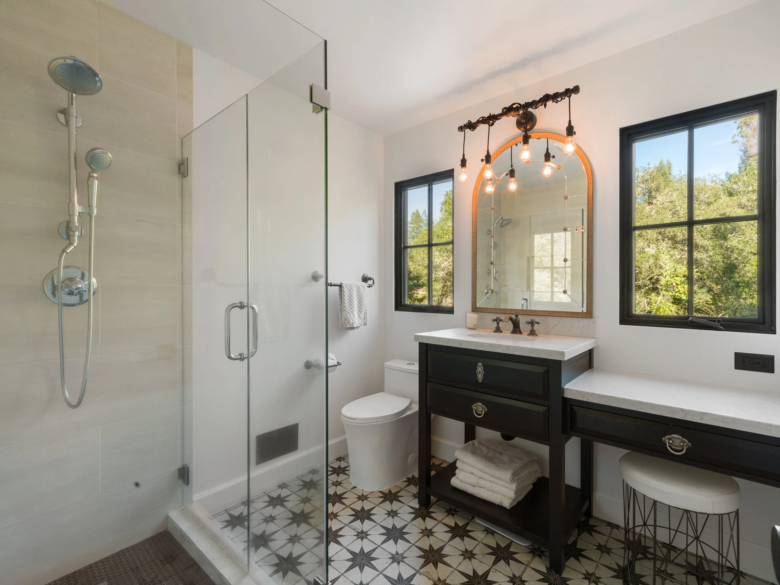 Modern bathroom with a walk-in shower, a toilet, a black vanity with a white marble countertop, a large window showing greenery outside, and decorative tile flooring. An arched mirror with lightbulbs hangs above the vanity.