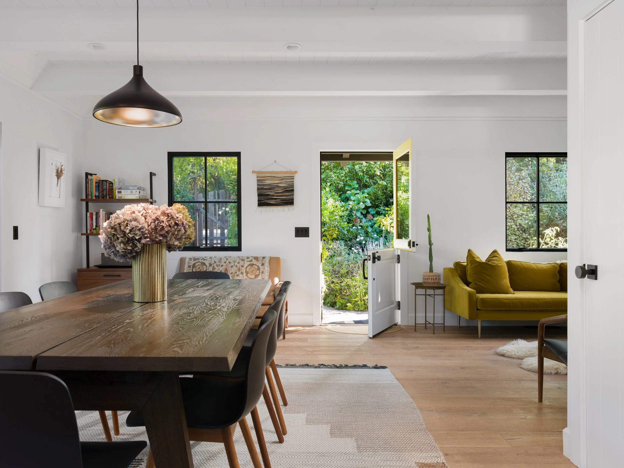 A dining area and living room with large windows. A wooden dining table with black chairs in the foreground. A vase of pink flowers on the table. A yellow sofa with pillows, a small side table, and a tall plant in the living room. Bright greenery vis