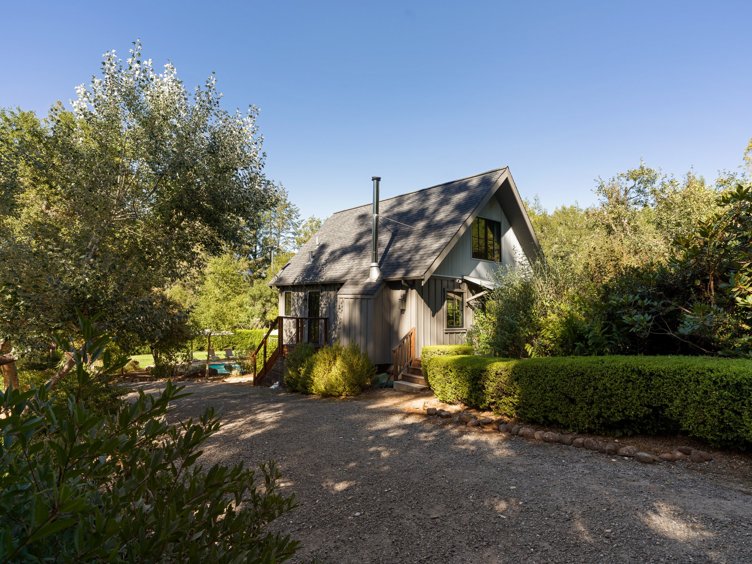 A house with a steep gable roof, surrounded by trees and shrubs, with a gravel driveway in front and a small deck with stairs.