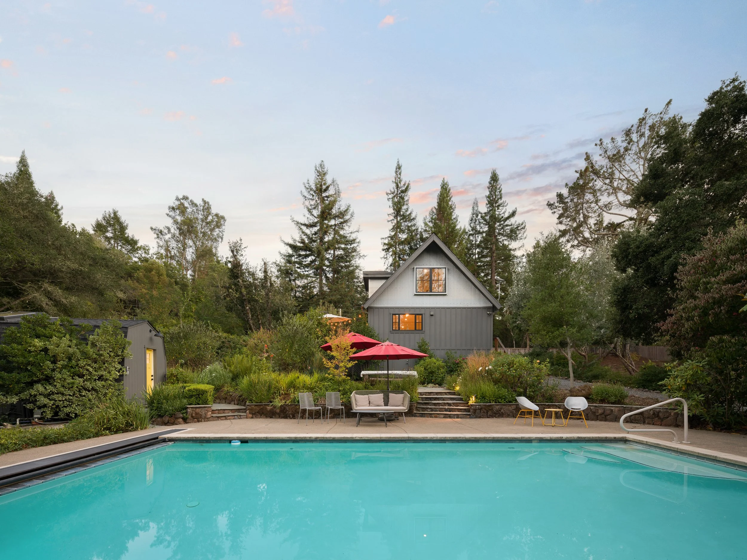 A backyard with a swimming pool, surrounded by outdoor furniture, red umbrellas, and garden beds. A small gray house with large windows is in the background, with tall trees and a partly cloudy sky behind it.