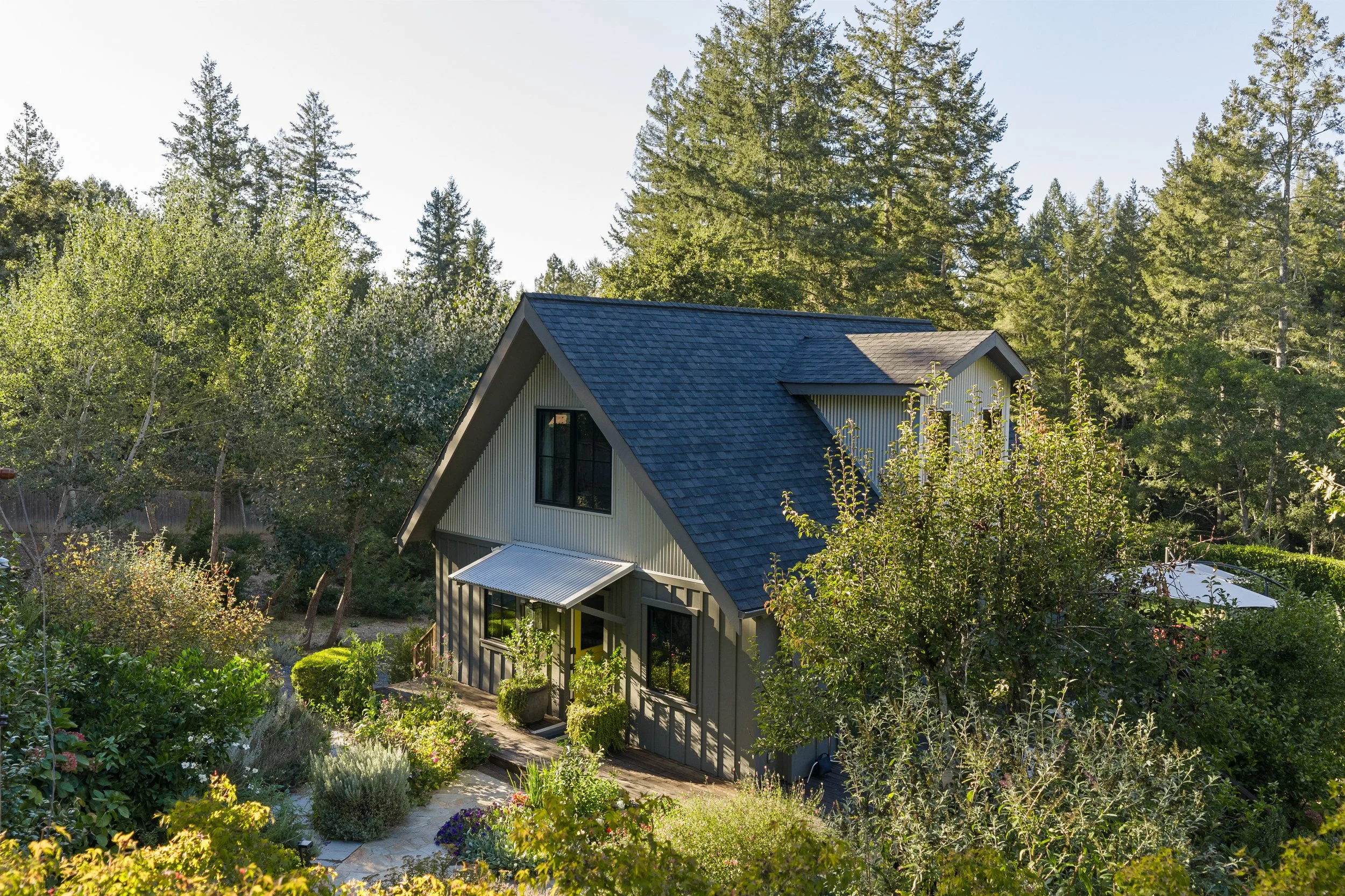 A house with a gray exterior and large windows, surrounded by lush greenery, trees, and a well-maintained garden on a sunny day.