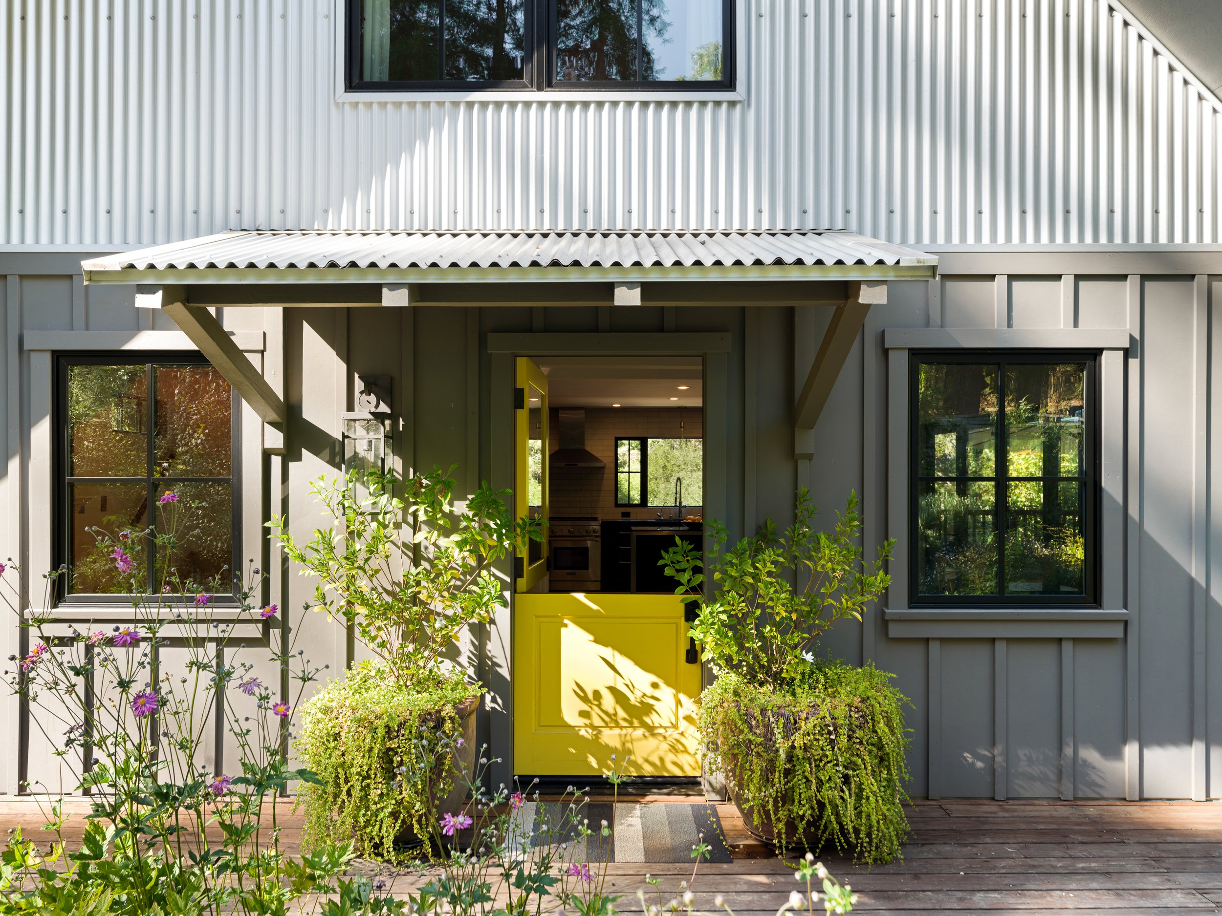 A house's gray exterior with large black-framed windows and a small porch or entryway with a bright yellow door, surrounded by green plants and purple flowers.