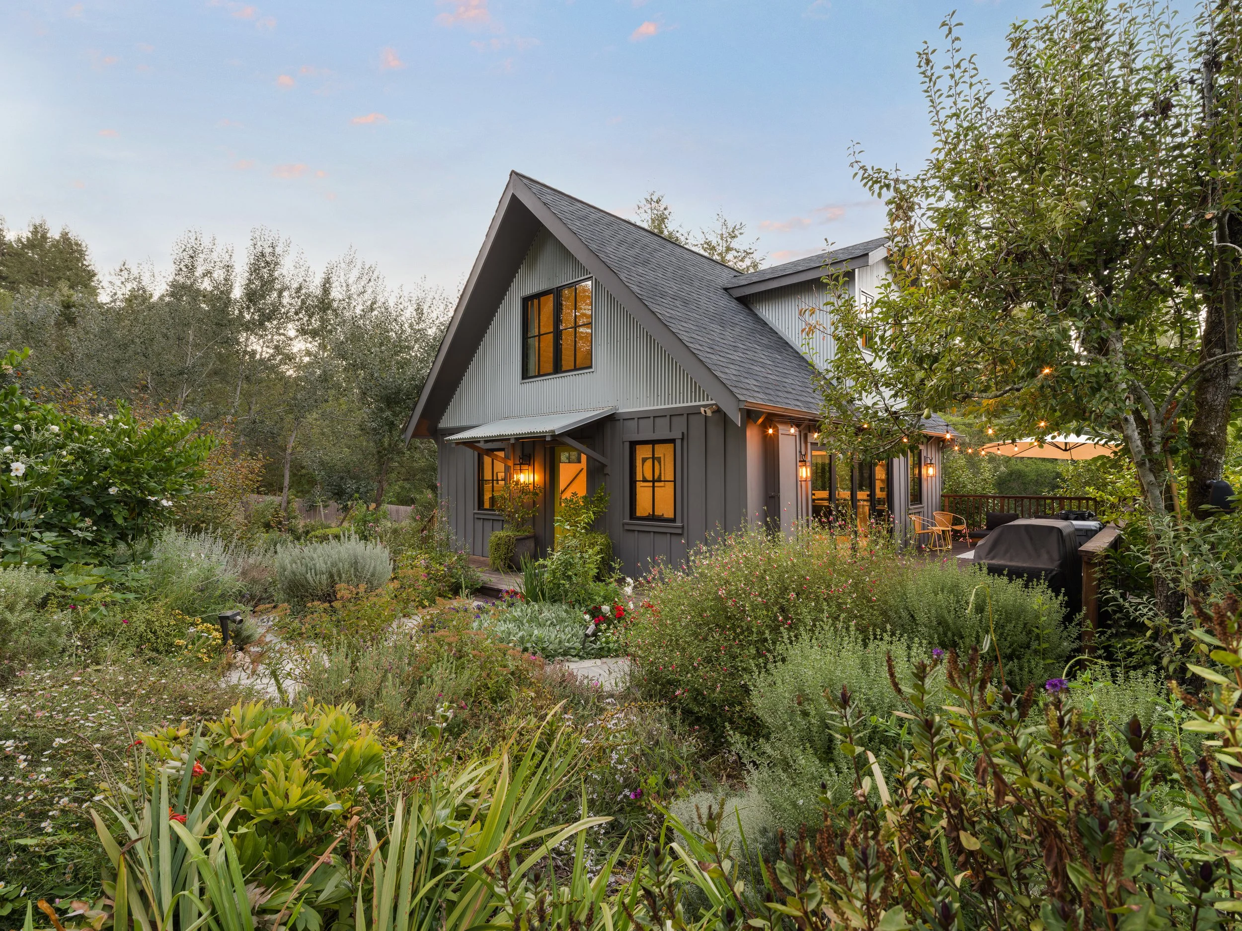 A gray house with black-framed windows and a sloped roof is surrounded by lush green plants and flowering bushes in a garden, with warm indoor lighting visible through the windows. There are string lights hanging on the side of the house, and an umbr