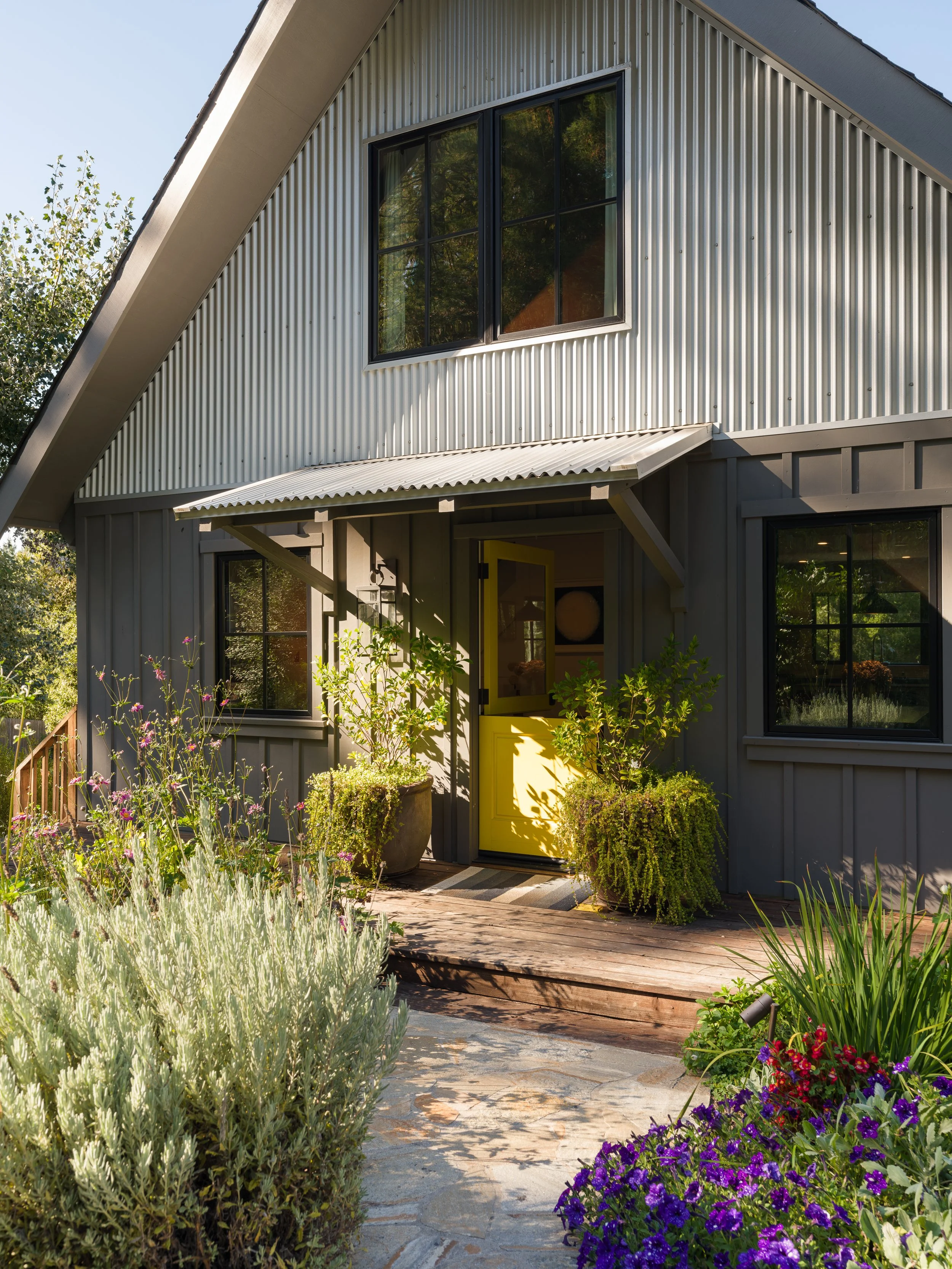 A house with gray metal siding, large windows, and a bright yellow front door, surrounded by lush greenery and colorful flowers in a garden.