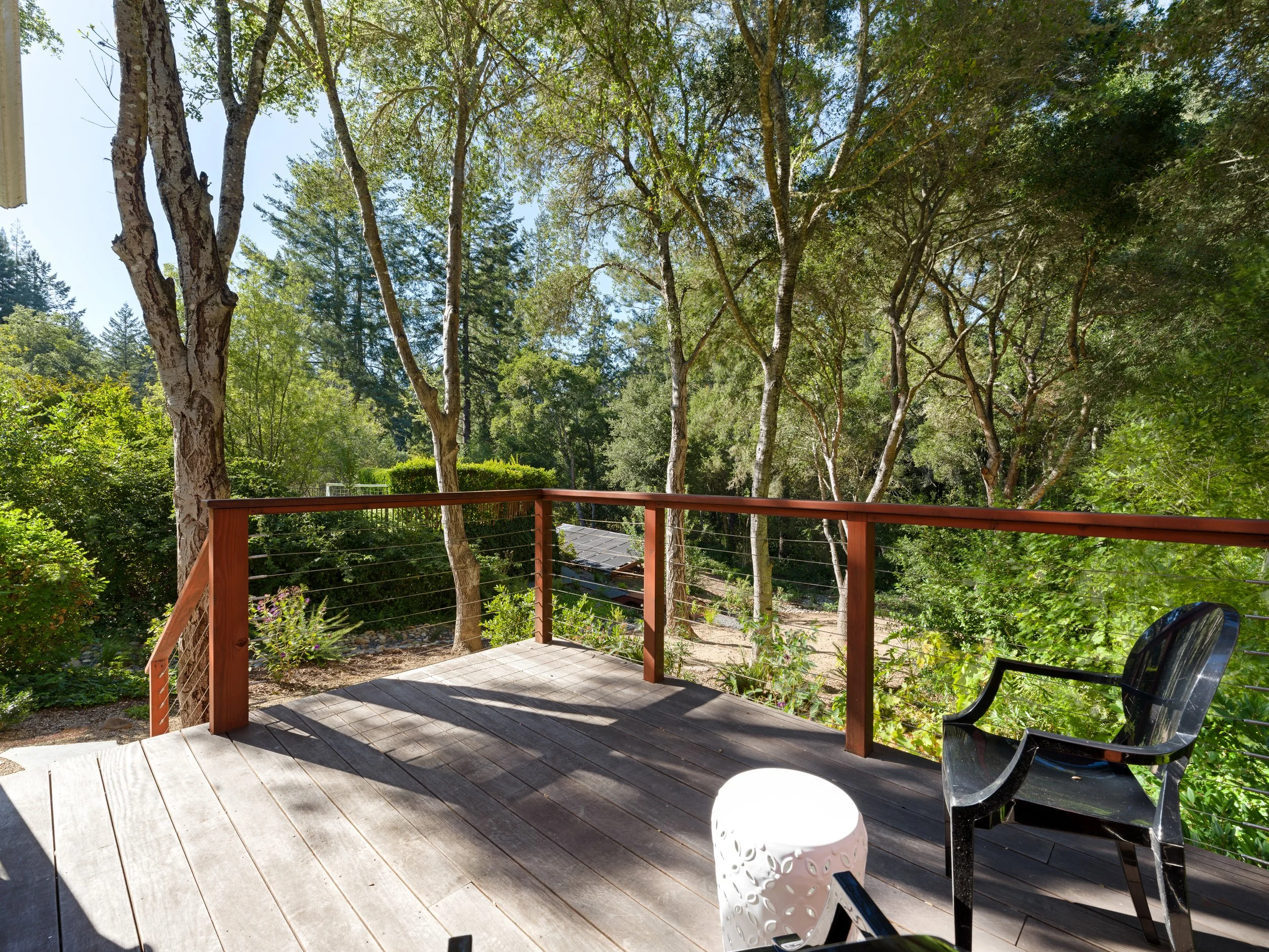 A backyard deck with wooden flooring, a black plastic chair on the right, a white table in the foreground, and tall trees with green leaves covering the sky beyond the deck.