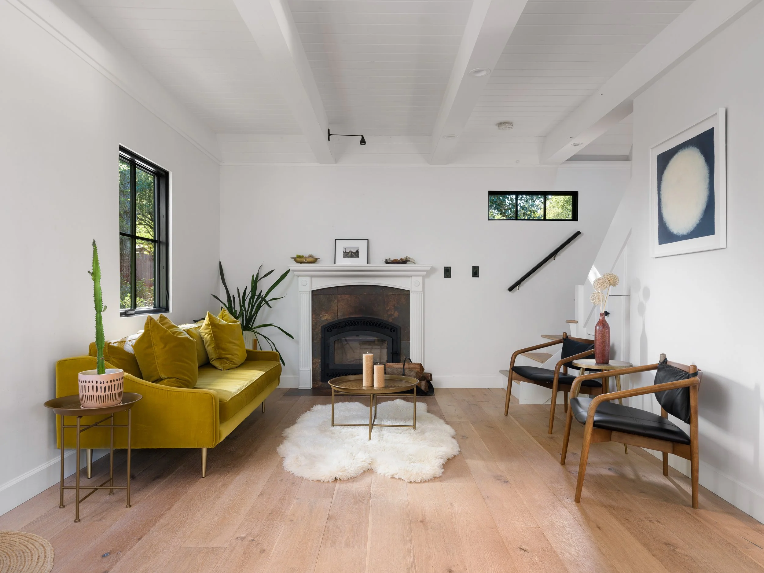Living room with white walls and ceiling, featuring a yellow sofa with pillows, a small round coffee table with candles, a fireplace with framed art above, large windows, a potted cactus, and modern chairs with a side table and vases.
